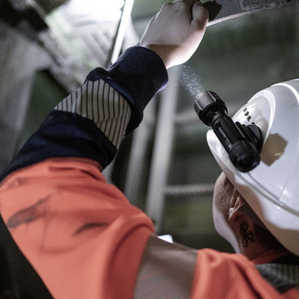 Osoba w pomarańczowym ubraniu roboczym i białym kasku świeci latarką. Drabina i belki konstrukcyjne znajdują się w tle.