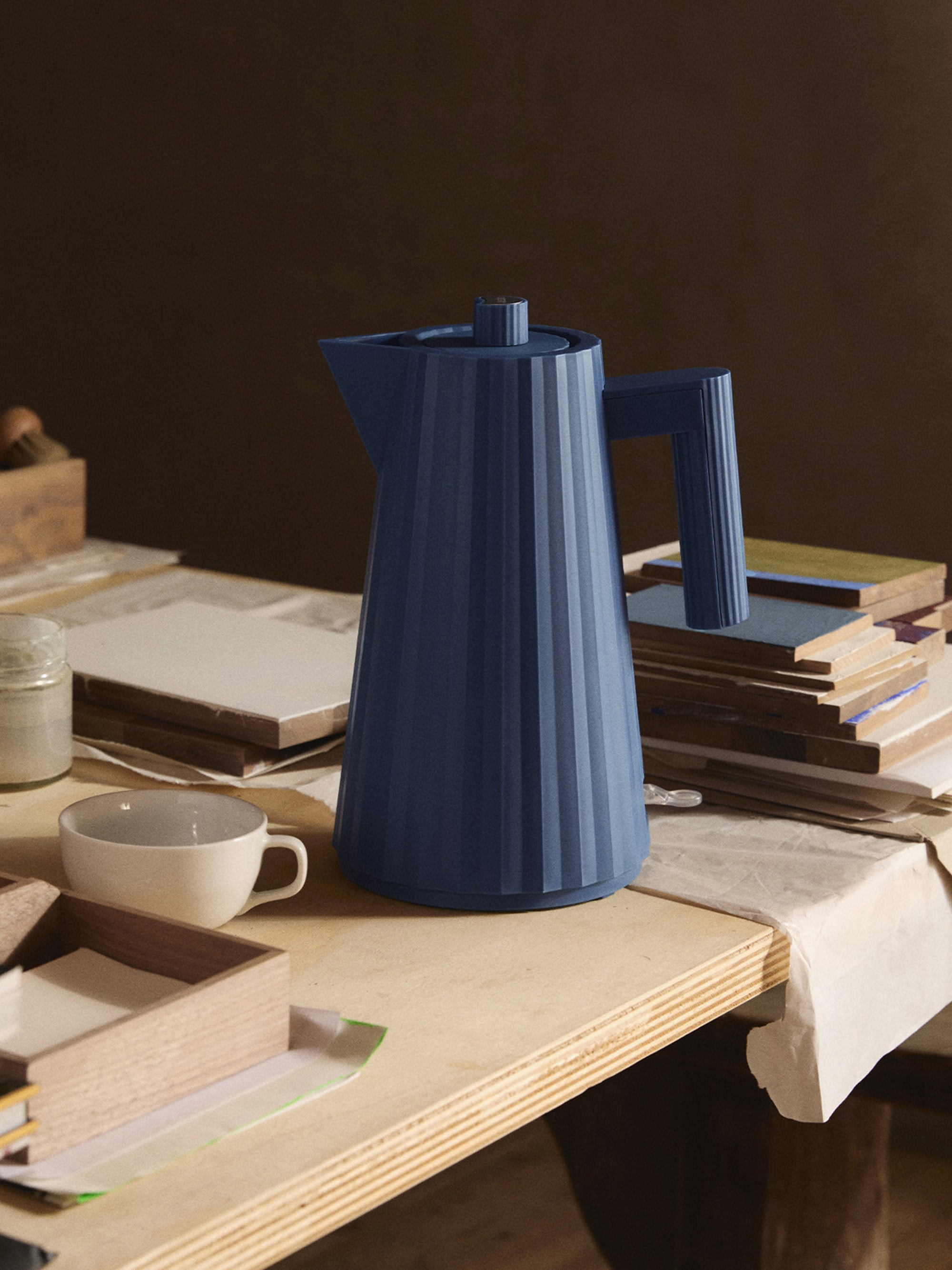 Una teiera blu a coste è su un tavolo di legno. Sono presenti anche una tazza bianca, una scatola di legno e dei libri.