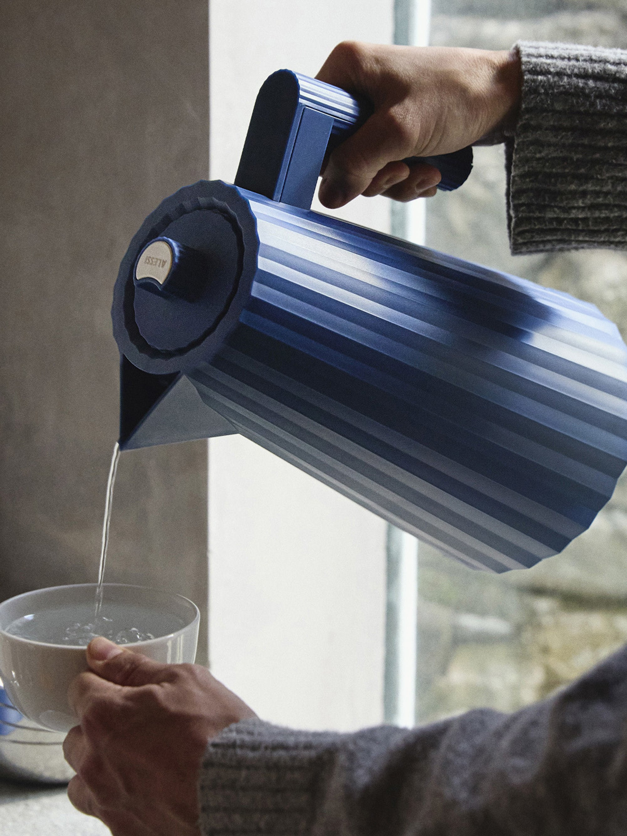 Una persona versa acqua da un bollitore blu in una tazza bianca. Maglione grigio e finestra visibili.