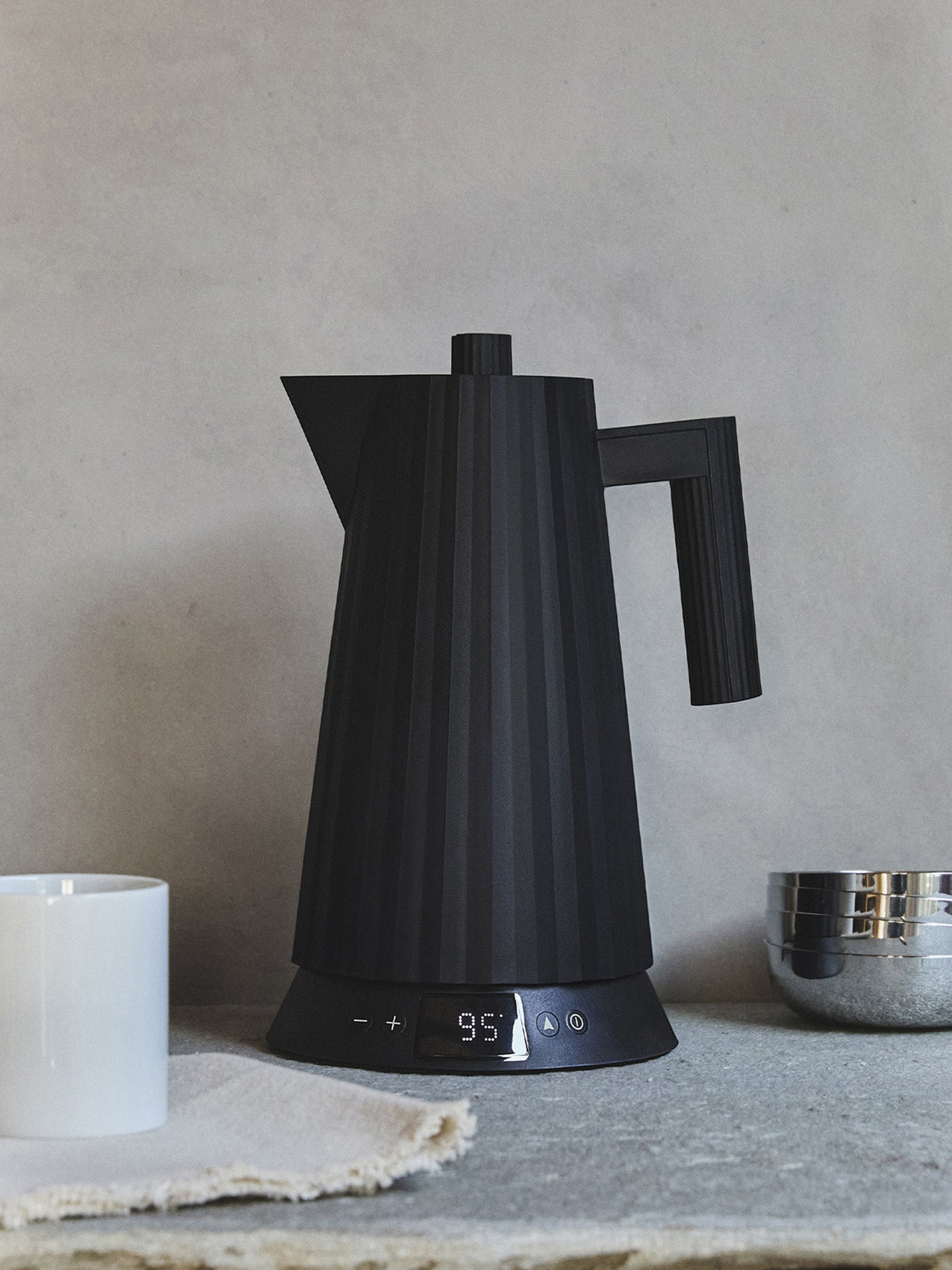 Un bollitore elettrico nero con display digitale, una tazza bianca e una ciotola in acciaio inossidabile su un piano di lavoro.