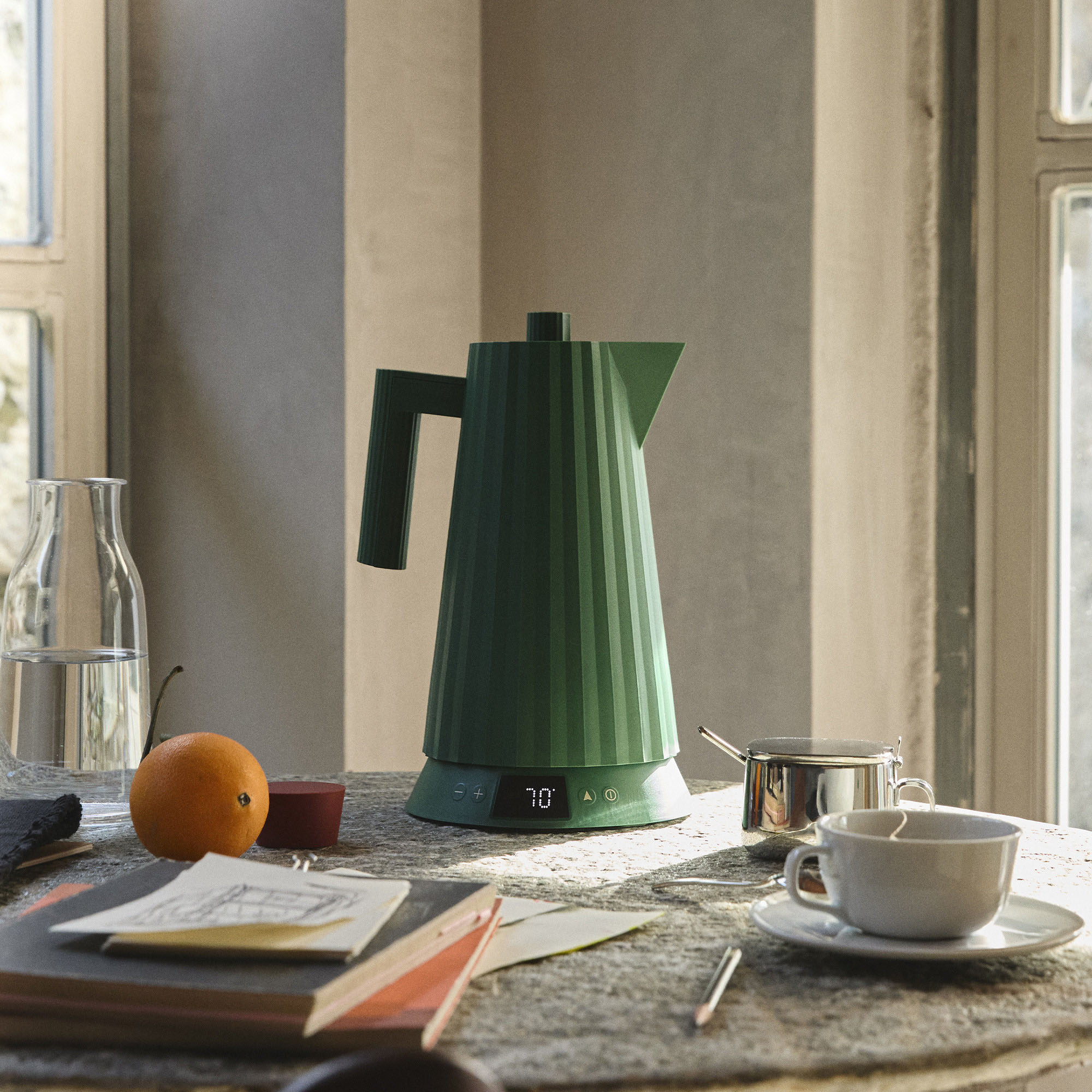 Un bollitore verde, tazza, arancia e bicchiere su un tavolo vicino a una finestra. La luce del sole illumina la scena.