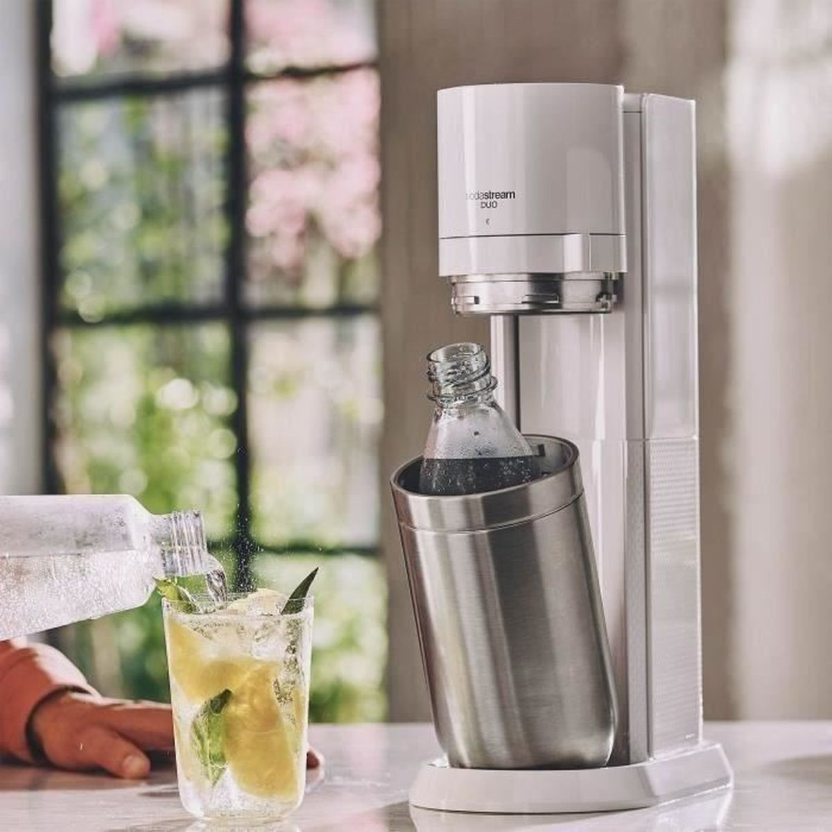 Eine weiße SodaStream Duo Maschine mit Glas und Flasche. Wasser wird in das Glas mit Zitrone und Eis gegossen.