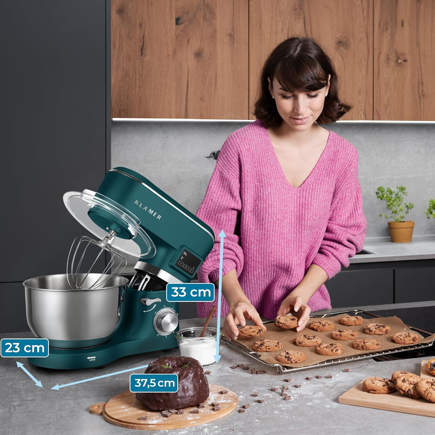 Eine Frau backt Kekse mit einem türkisfarbenen Mixer. Kekse befinden sich auf einem Tablett und Holzbrettern. Schokochips und ein Kuchen sind ebenfalls vorhanden.
