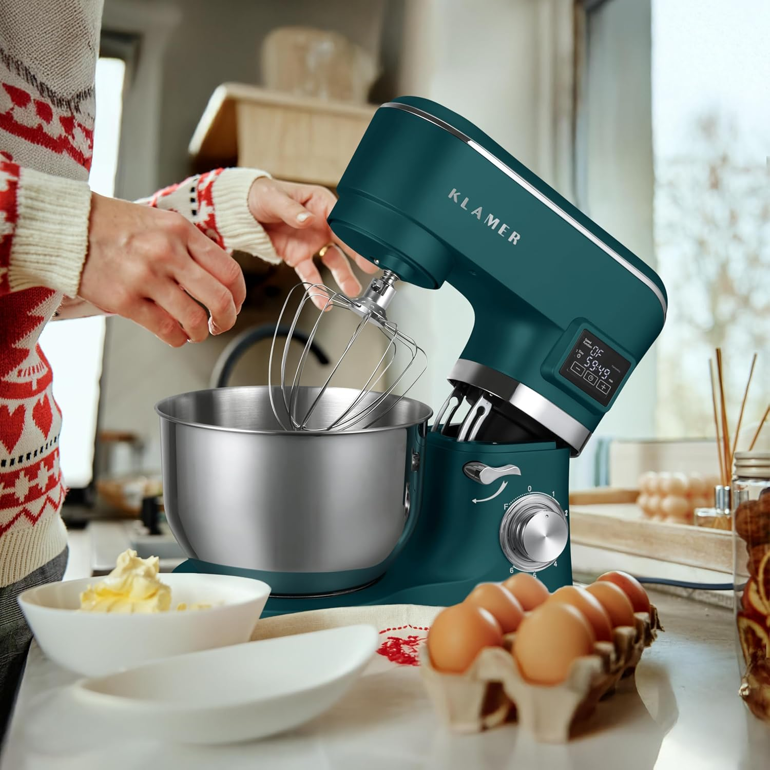 Eine Person benutzt einen türkisfarbenen Küchenmixer mit Schneebesenaufsatz. Eier, Butter und Schüsseln stehen auf der Theke.