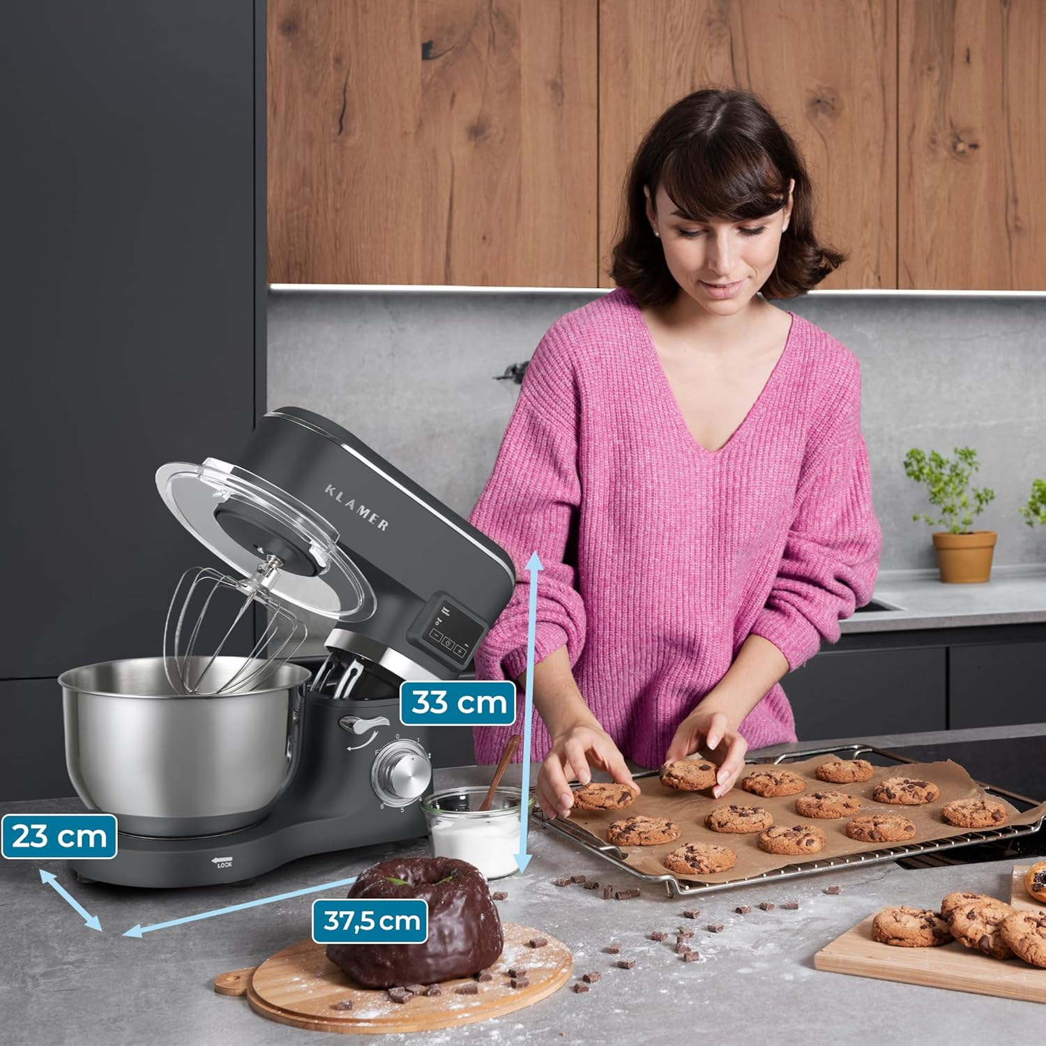 Frau im rosa Pullover mit Keksen. Mixer im Hintergrund. Schokoladenkuchen und Zutaten auf dem Tisch. Küchenumgebung.