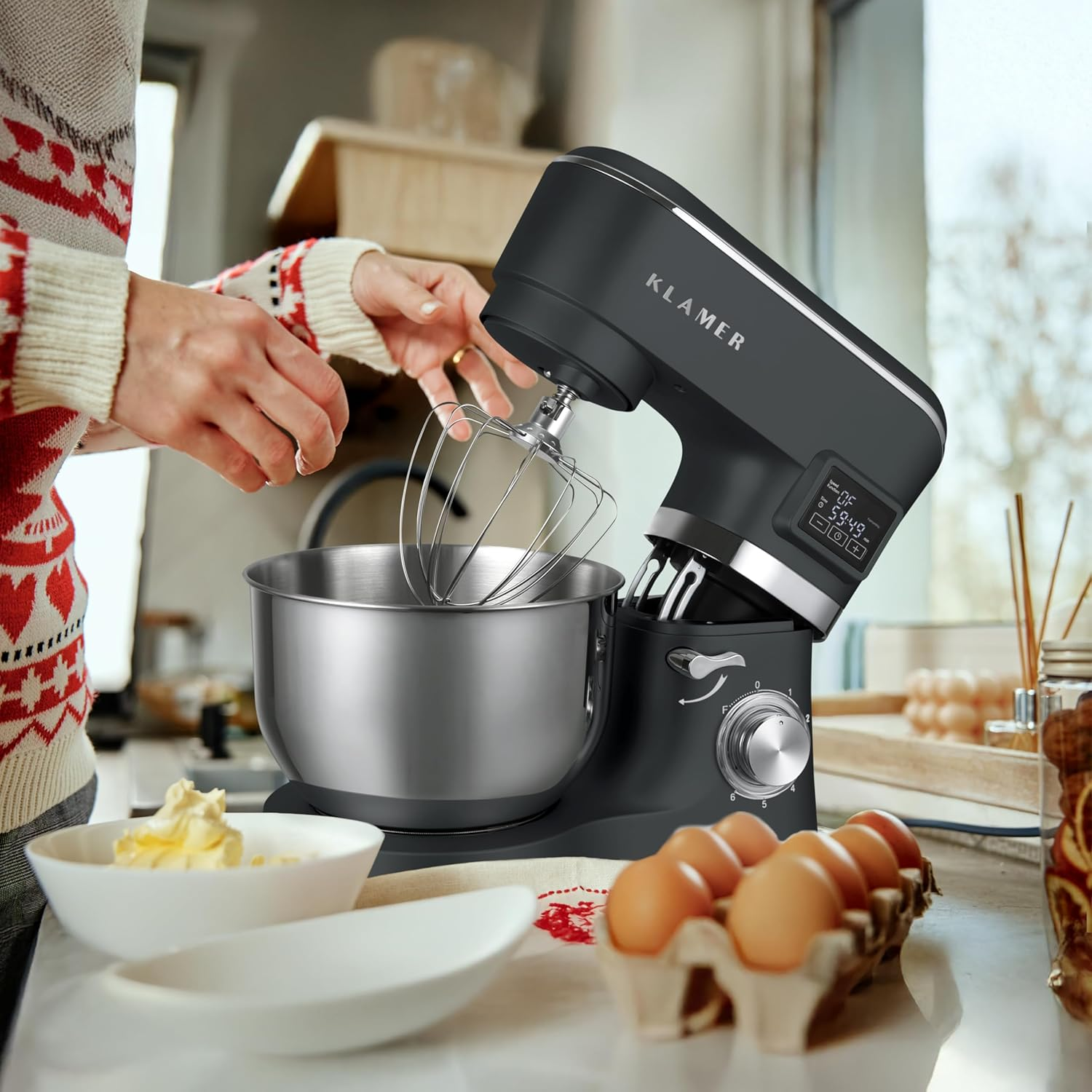Eine Person benutzt eine schwarze Klamer-Küchenmaschine. Auf der Theke stehen Eier, Butter und Schüsseln. Die Person trägt einen gemusterten Pullover.