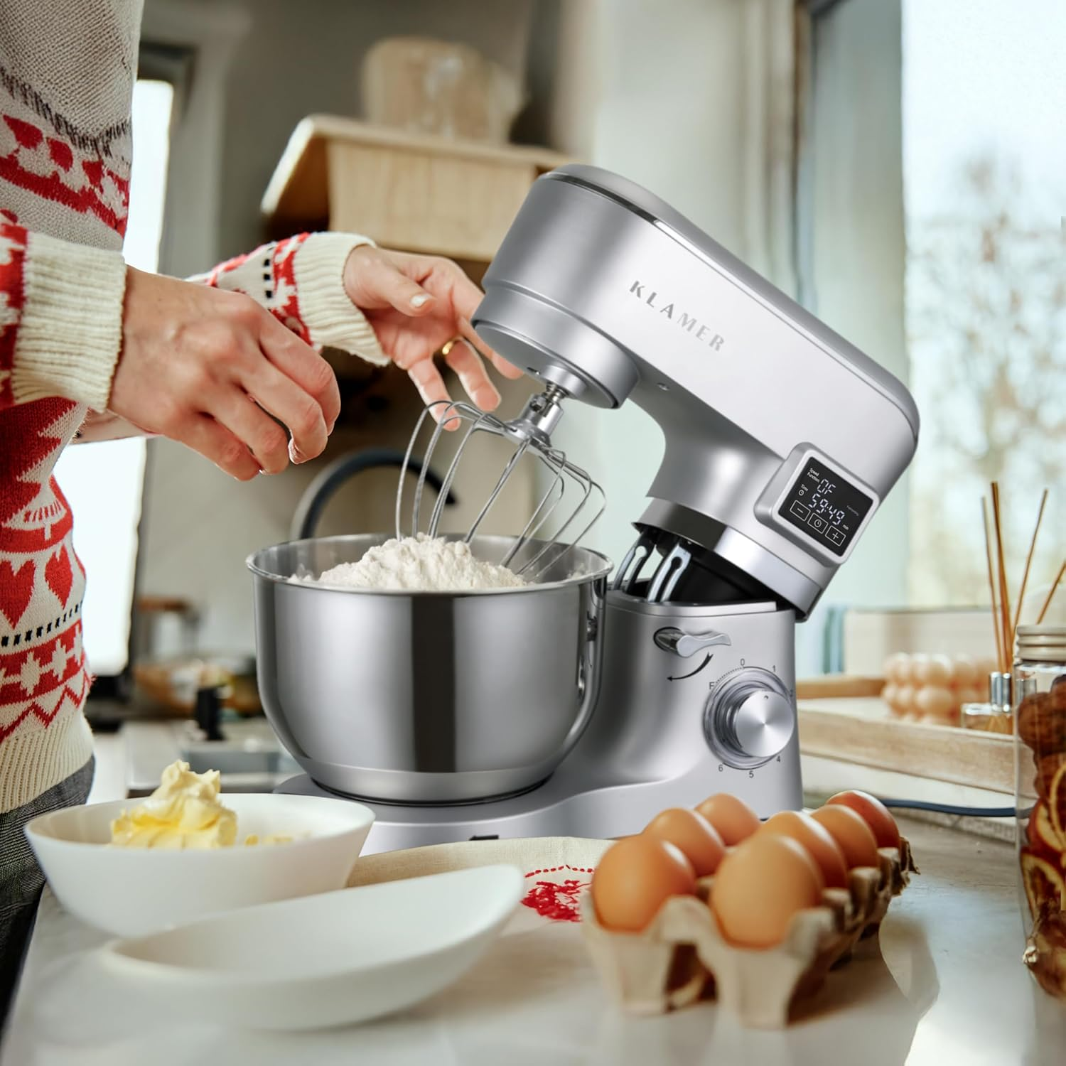 Eine Person verwendet einen silbernen Küchenmixer, um Mehl in einer Schüssel zu verrühren. Eier und Butter liegen auf dem Tisch. Die Person trägt einen gemusterten Pullover.