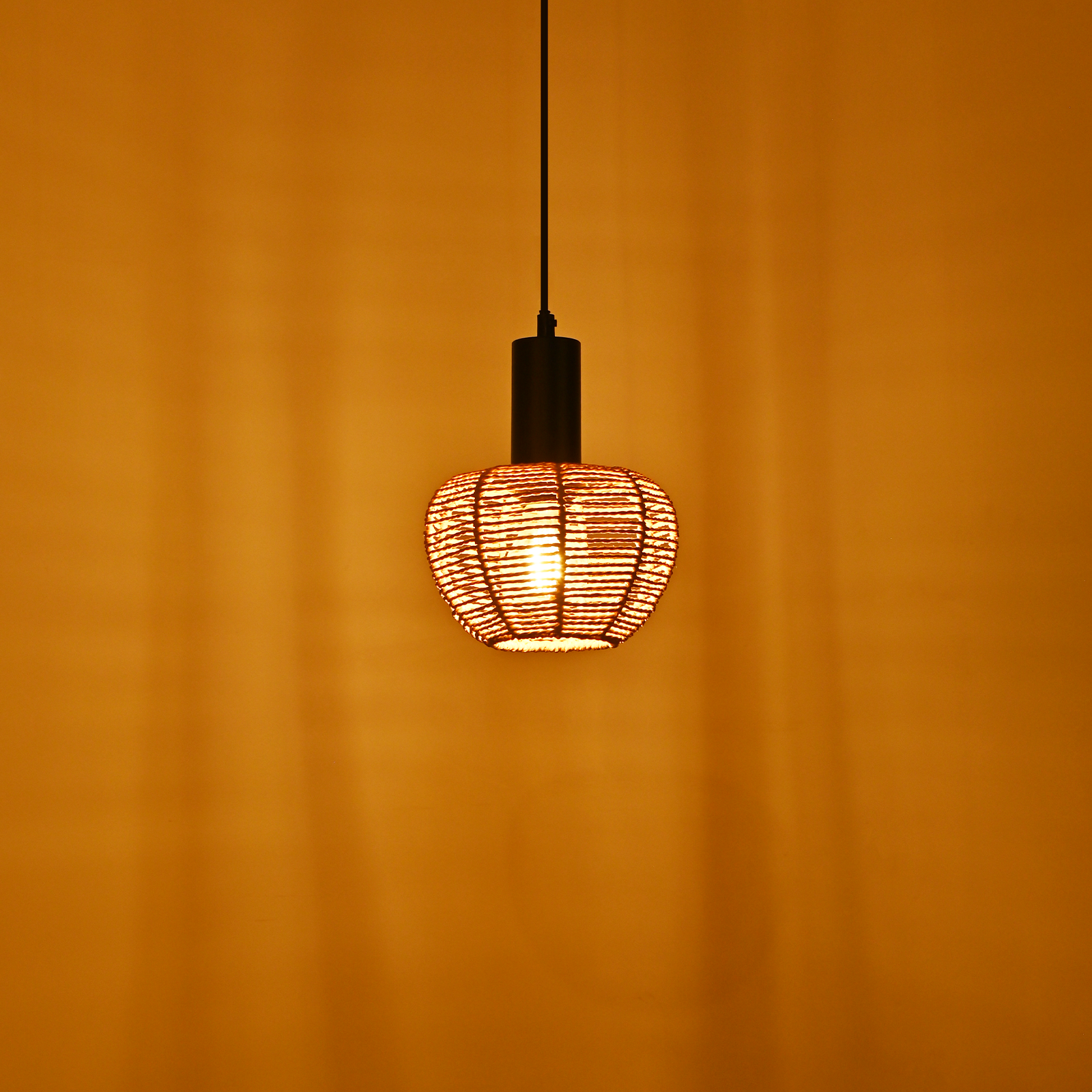 Lampa wisząca wisi na żółtej ścianie. Lampa ma tkany klosz z widoczną żarówką.