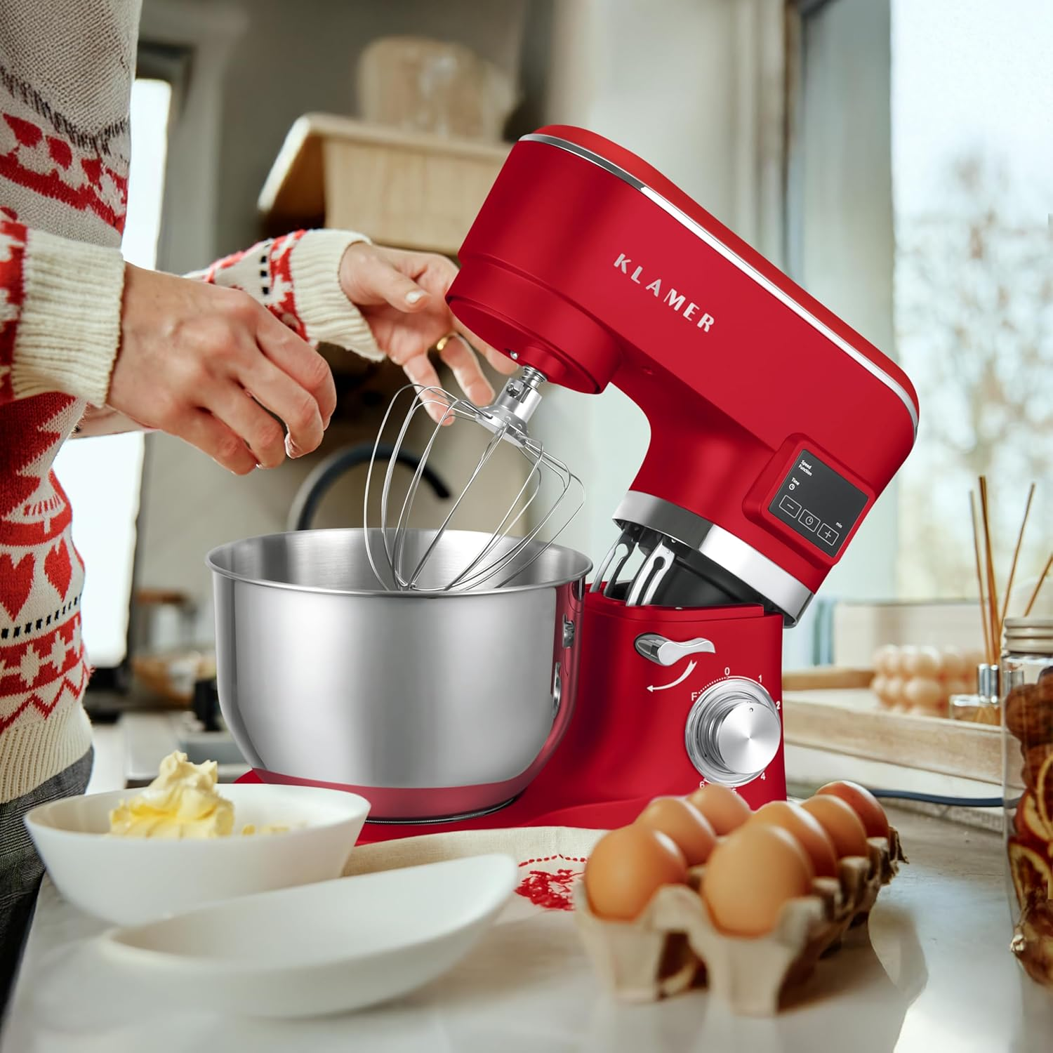Ein roter Küchenmixer mit Edelstahlschüssel, Eiern und einer Frau, die backen will. Zutaten sind auf dem Tisch platziert.