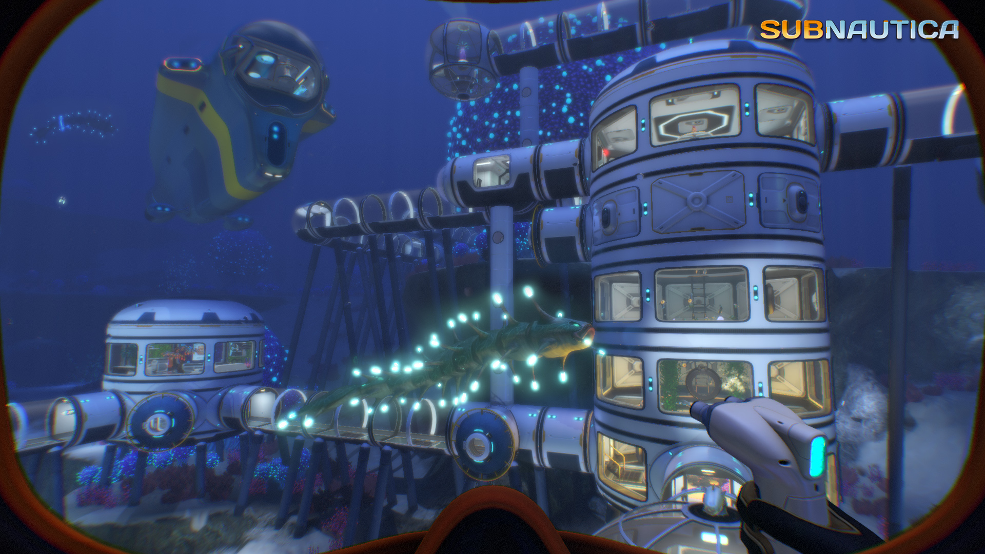 Podwodna baza z pojazdem i stworzeniem. Ręka trzymająca narzędzie jest na pierwszym planie. Widoczne jest logo Subnautica.