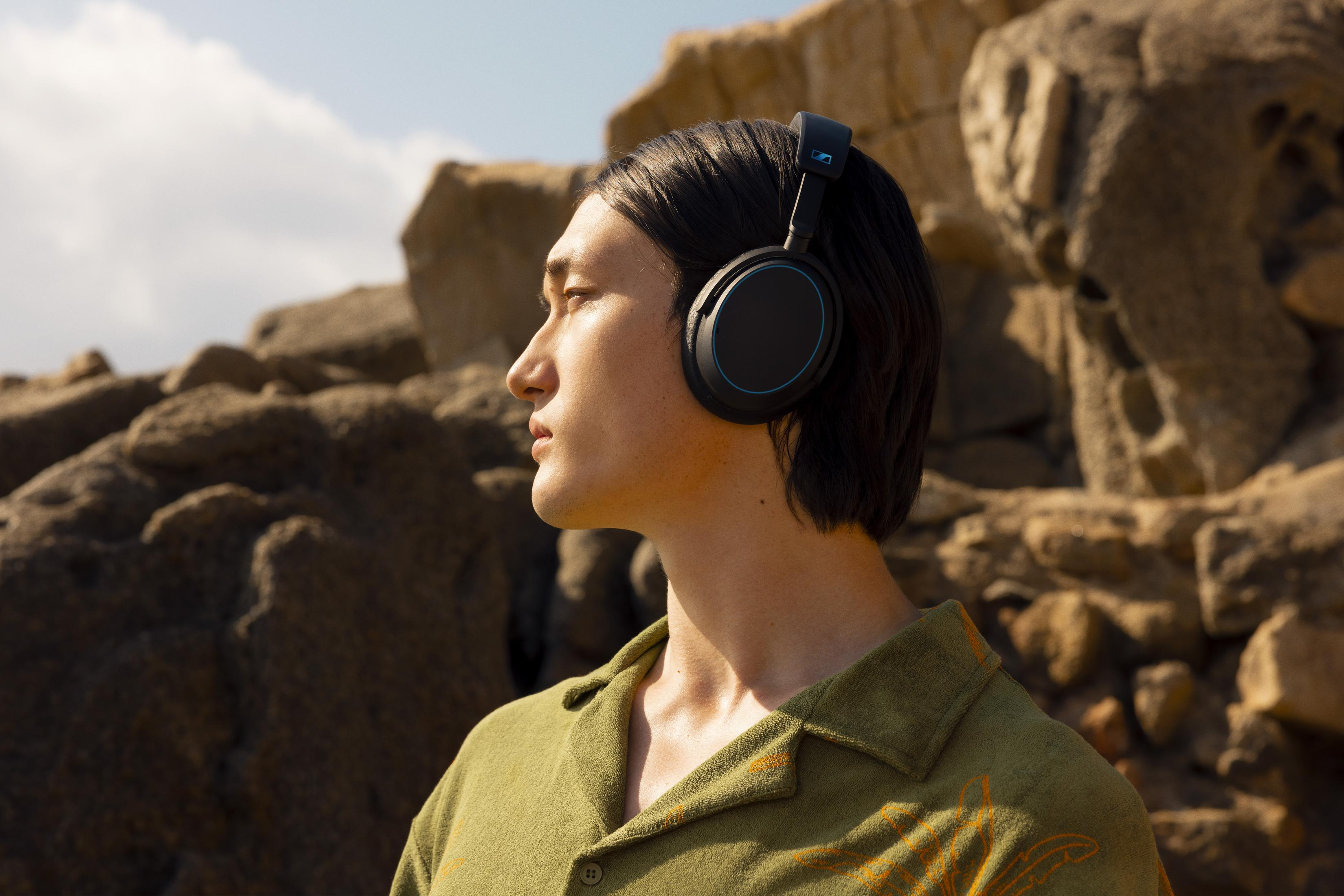 Eine Person mit Kopfhörern blickt weg, vor einem felsigen Hintergrund und einem Himmel mit Wolken. Die Person trägt ein grünes Hemd.