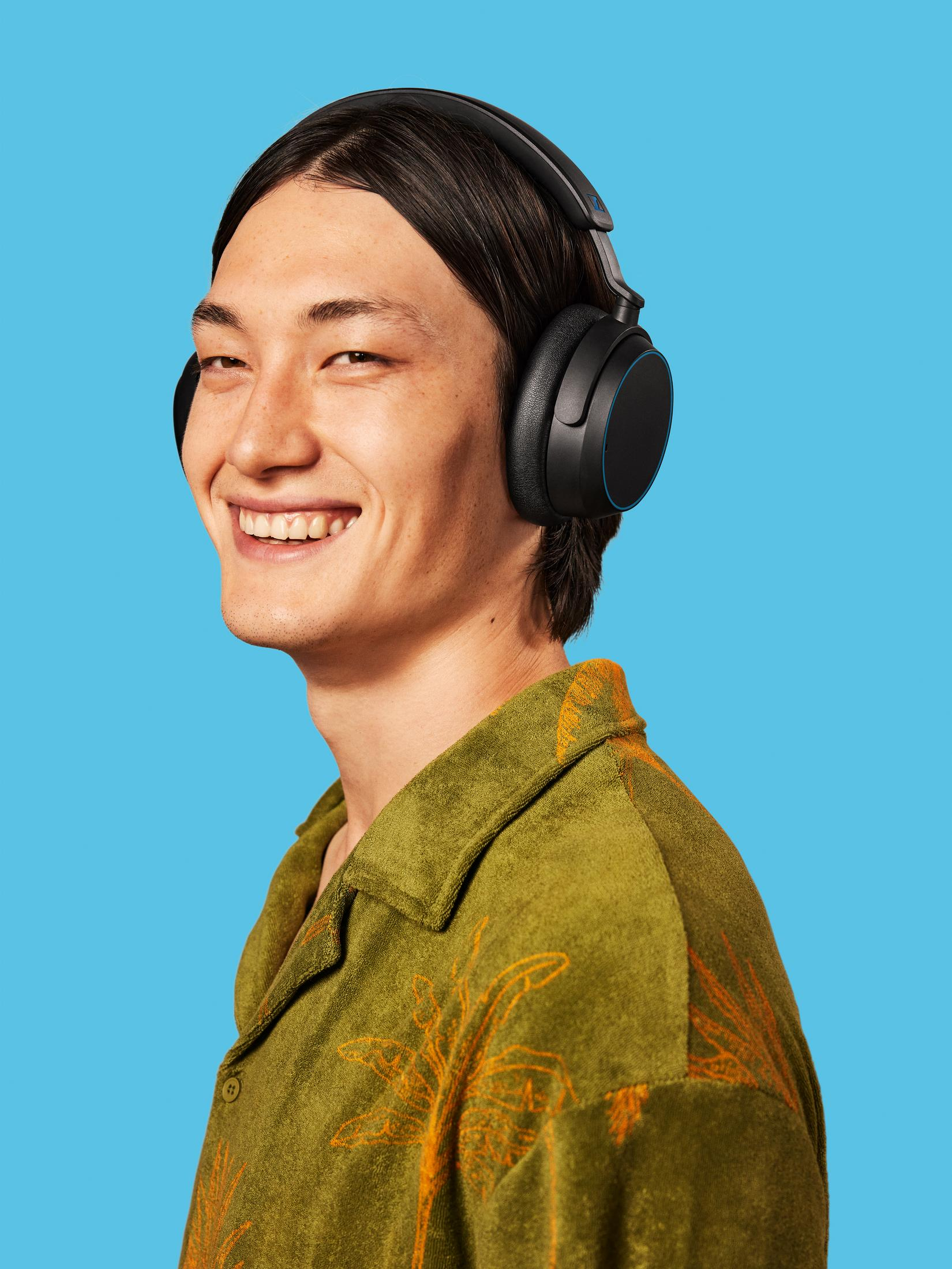 Ein lächelnder Mann mit Kopfhörern vor blauem Hintergrund, trägt ein grünes Hemd mit orangen Mustern.
