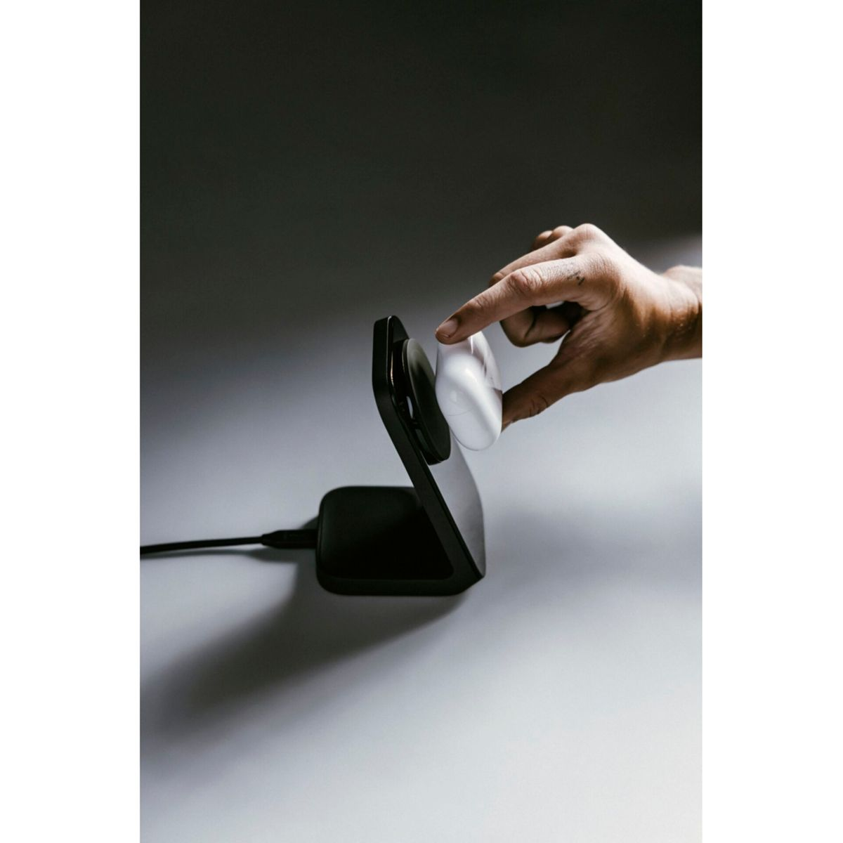 Eine Hand hält einen weißen Ohrhörer in der Nähe eines schwarzen Ladeständers. Der Ständer steht auf einer weißen Oberfläche mit dunklem Hintergrund und Kabel.