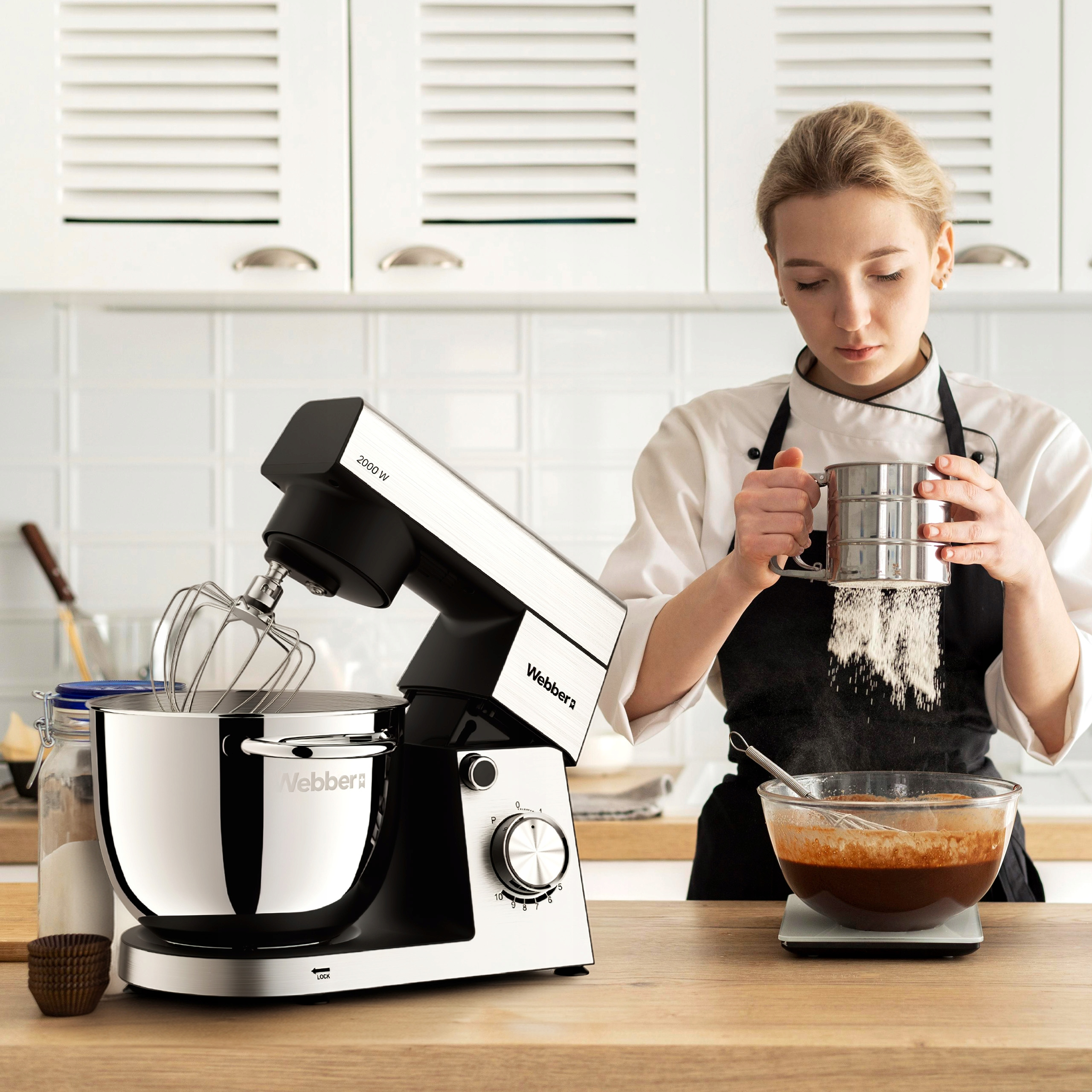 Kobieta przesiewa mąkę do miski za pomocą sita, podczas gdy mikser i składniki stoją na drewnianym blacie.