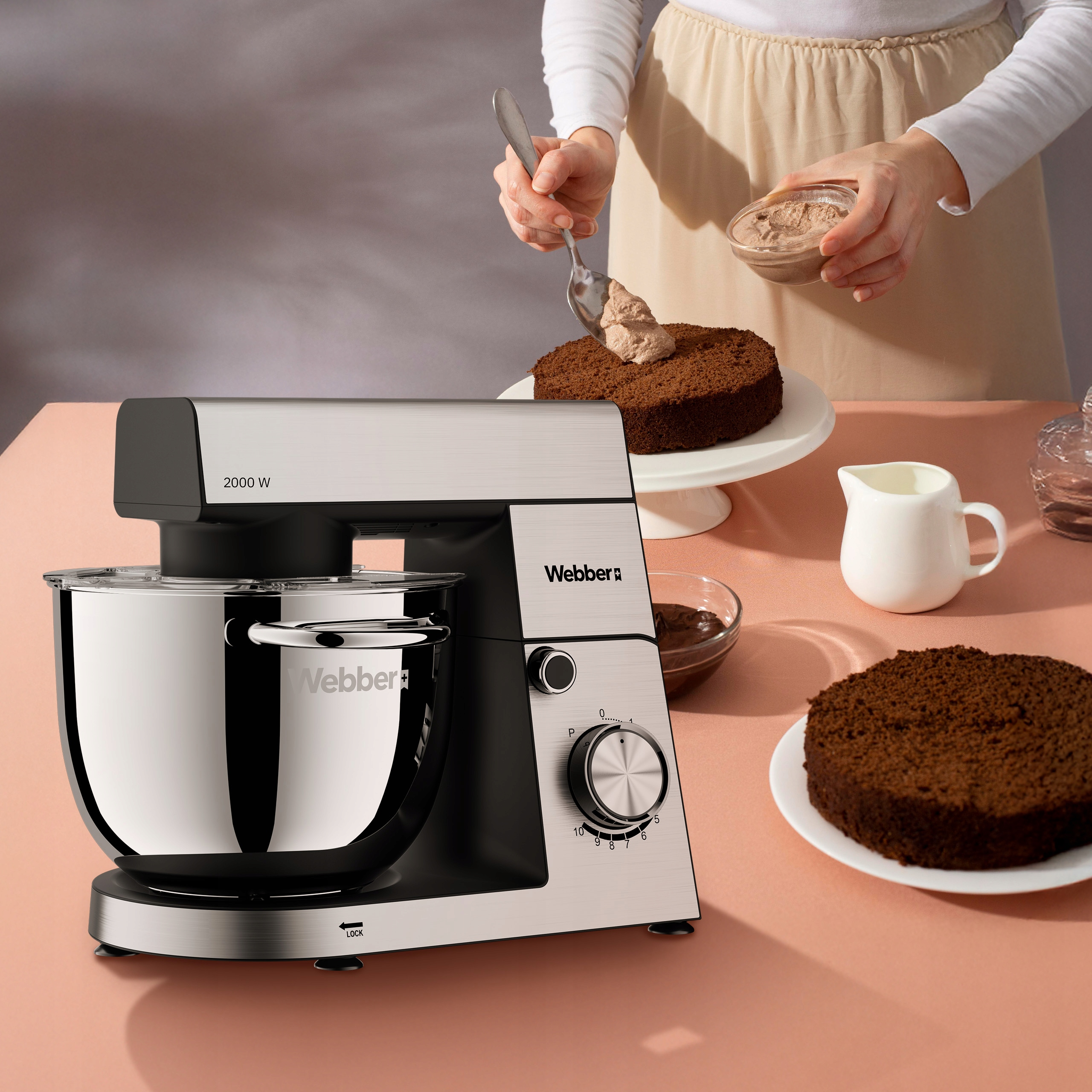 Scena kuchenna z mikserem do ciasta, ciastem lukrowanym i kawałkami ciasta na różowej powierzchni.