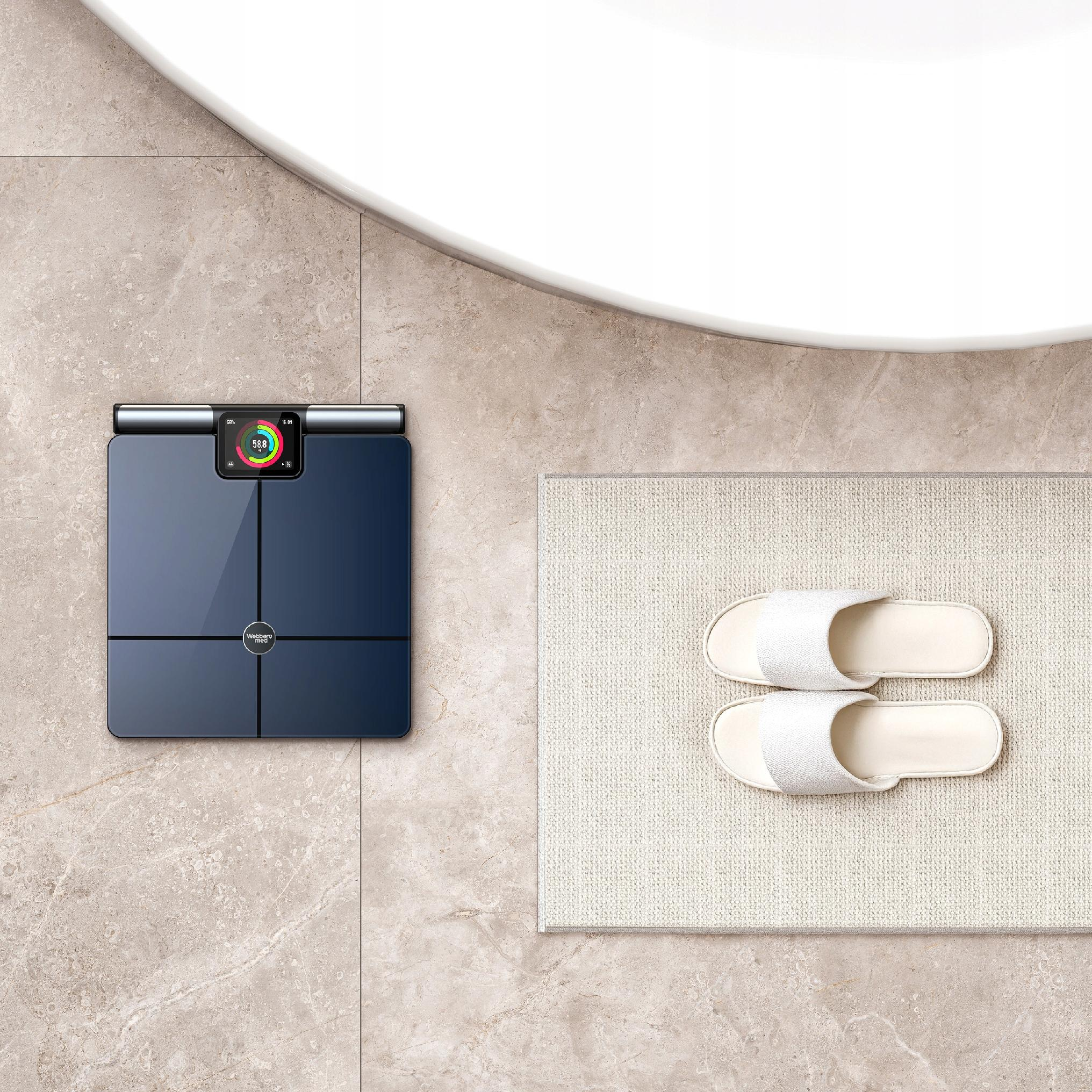 Scena łazienkowa z wagą cyfrową, białymi kapciami na dywaniku i zakrzywioną umywalką, wszystko na kafelkach.