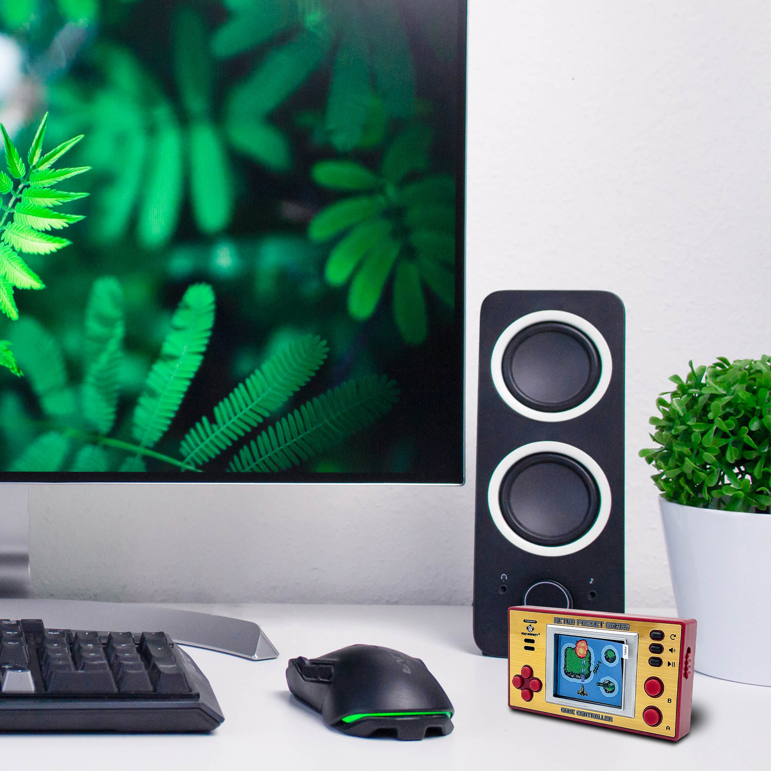 Ein Schreibtisch-Setup mit einem Monitor, der grüne Blätter zeigt, einem Lautsprecher, einer Handheld-Spielkonsole, Maus, Tastatur und einer Pflanze.