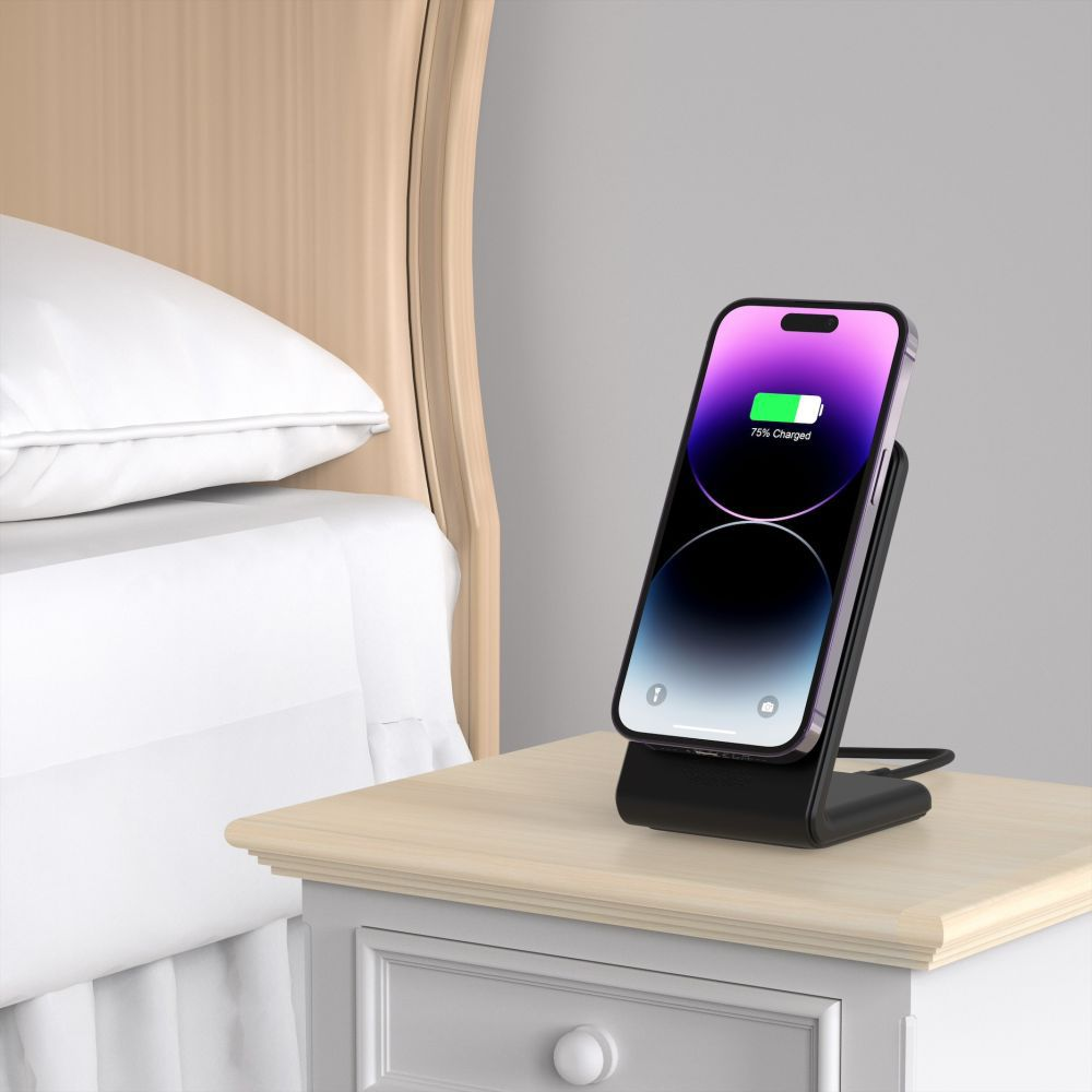 Un téléphone en charge sur un support. L'écran du téléphone affiche 75% de charge. A côté du téléphone, une table de chevet avec un lit et une tête de lit.