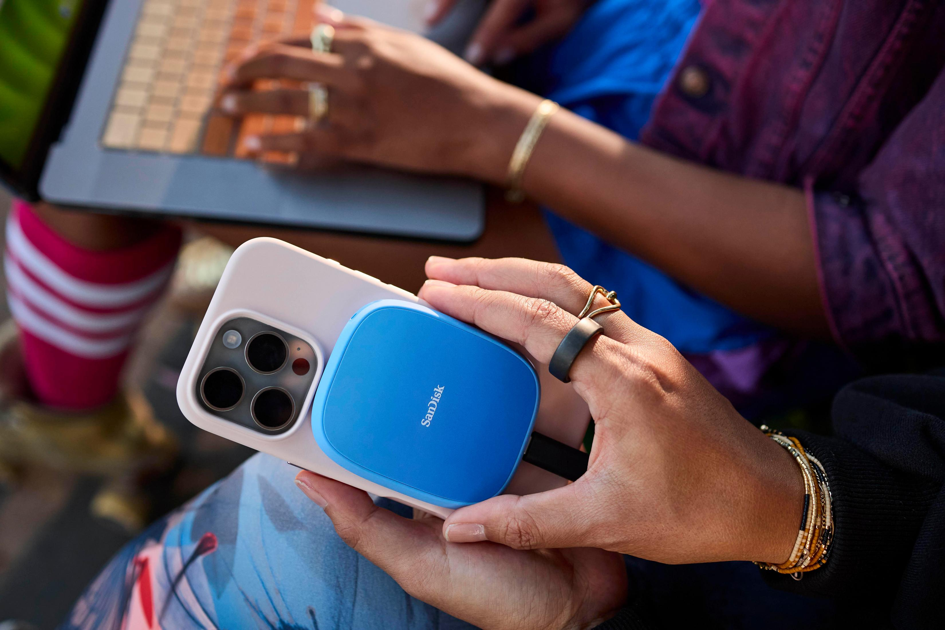 Una persona tiene in mano un telefono con un caricatore Sandisk blu. Un'altra persona lavora su un laptop.