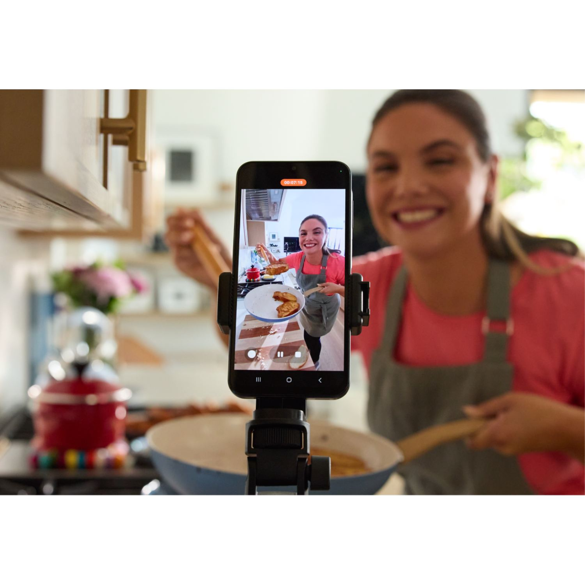 Kobieta uśmiecha się, filmując siebie podczas gotowania. Telefon na statywie. Kuchnia w tle z patelniami, kuchenką i czerwonym czajnikiem.