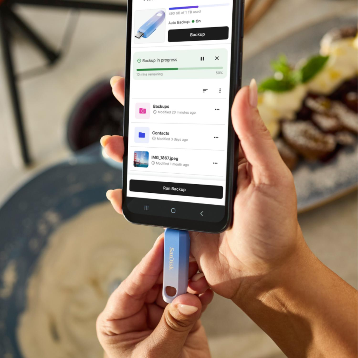 Ręka trzyma telefon z trwającą kopią zapasową. Niebiesko-biały pendrive jest trzymany pod telefonem. Jedzenie w tle.