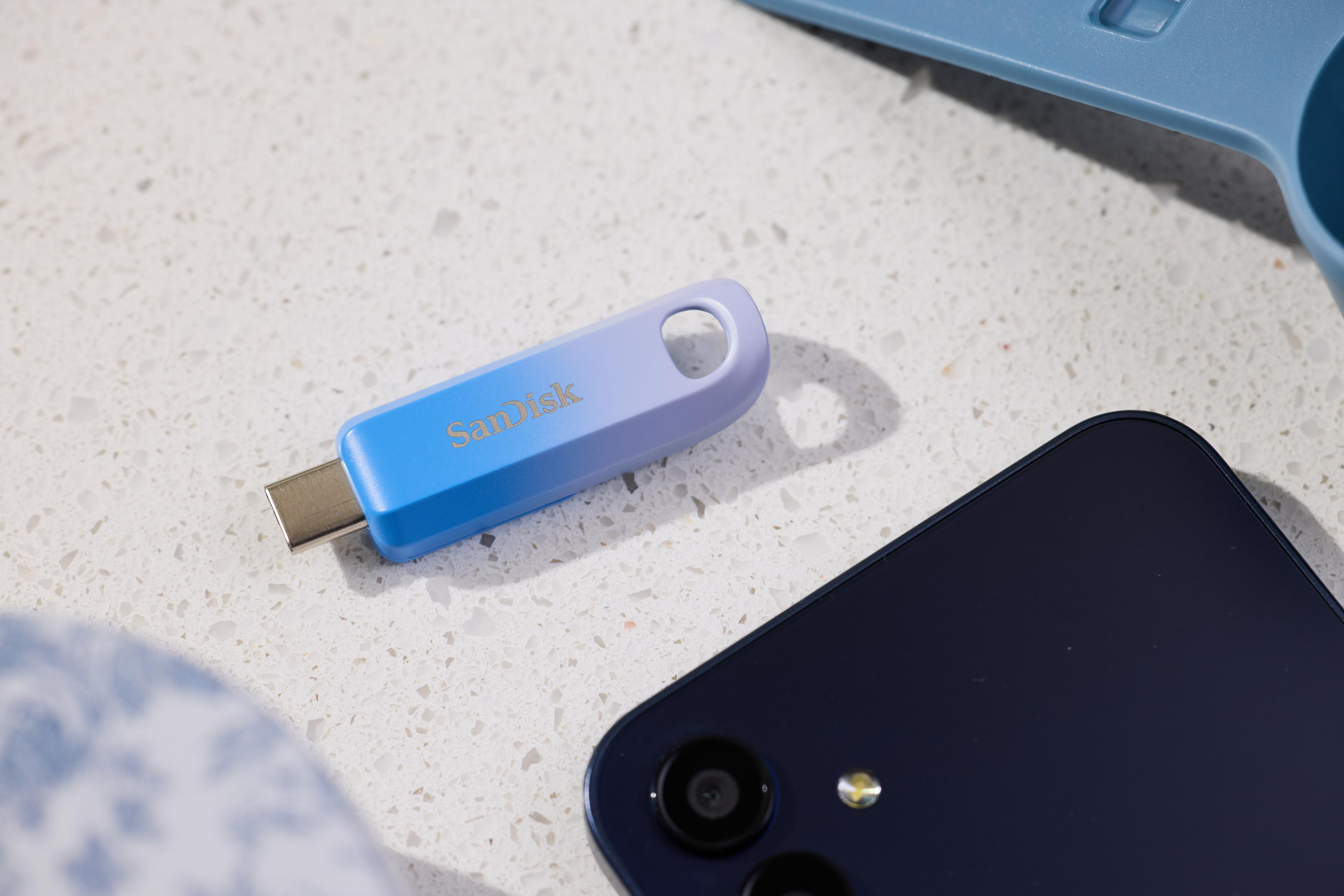 Niebiesko-fioletowy pendrive SanDisk leży na białej powierzchni obok granatowego telefonu.