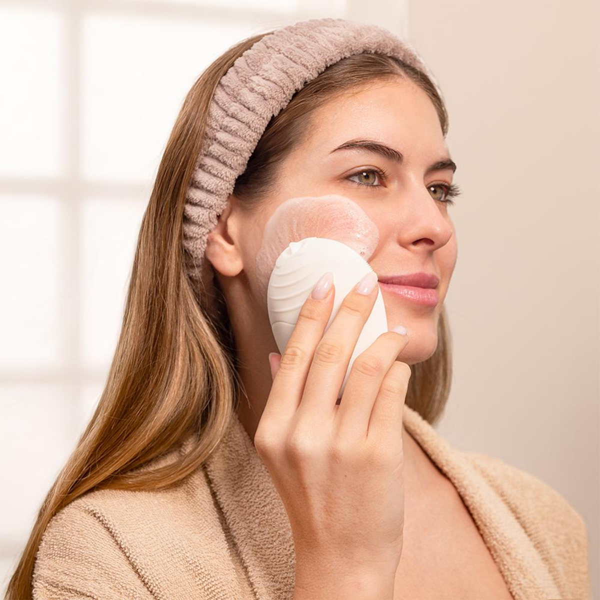 Una donna usa un dispositivo di pulizia viso bianco sulla guancia. Indossa una fascia e un accappatoio. Crema rosa sul viso.
