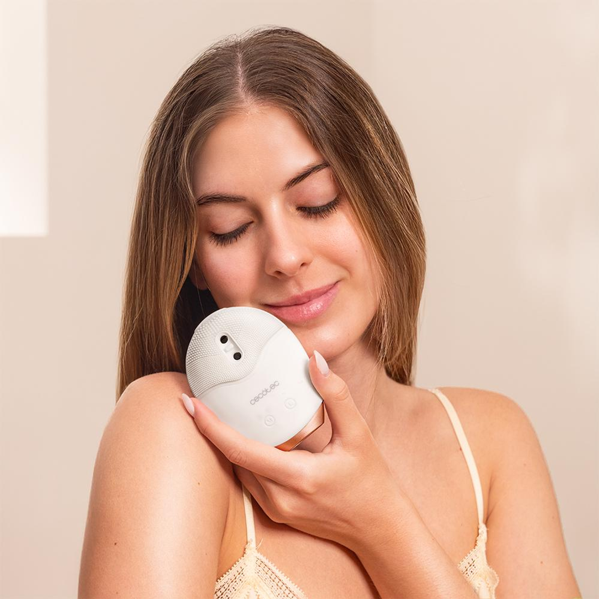 Una donna tiene un dispositivo bianco per la pulizia del viso contro la spalla, con gli occhi chiusi. Sorride. Lo sfondo è una parete beige.