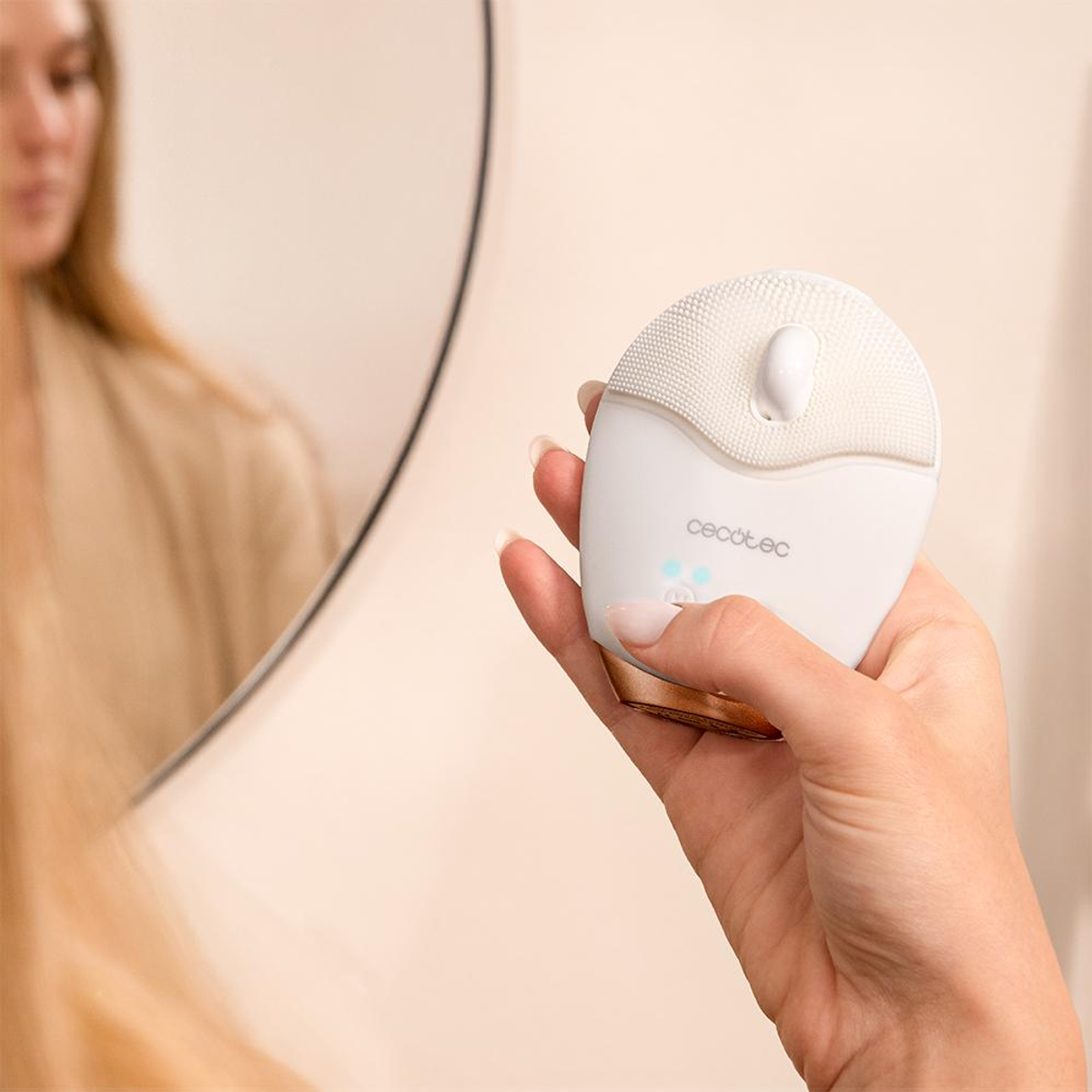 Una donna tiene in mano un dispositivo bianco per la pulizia del viso davanti a uno specchio. Il dispositivo ha una forma arrotondata con la parte superiore strutturata.
