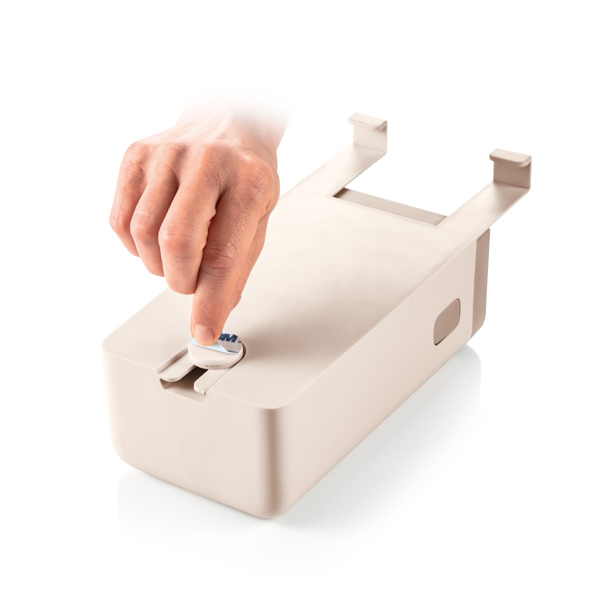 Eine Hand drückt einen kleinen, runden Klebstoff auf ein beiges rechteckiges Objekt mit zwei oberen Haken. Der Hintergrund ist weiß.