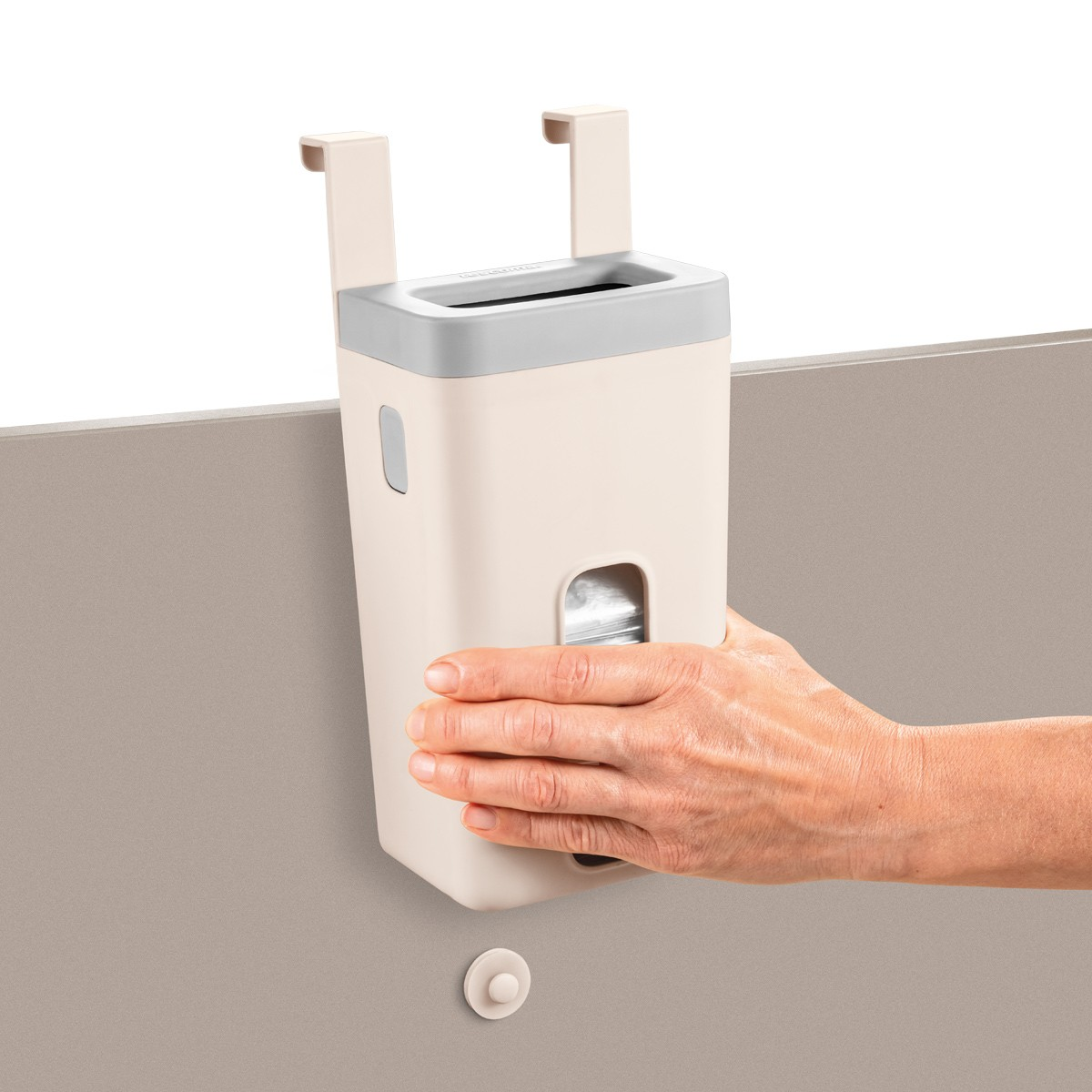 Ein beiger Hänge-Mülleimer mit grauem Deckel wird von einer Hand gehalten. Er hängt über einer braunen Oberfläche, einer weißen Wand und einem weißen Haken.