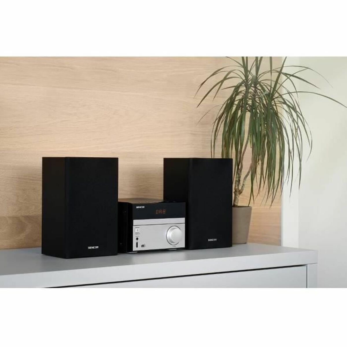 Un impianto stereo con altoparlanti neri, pannello di controllo argentato e pianta in vaso su superficie bianca.