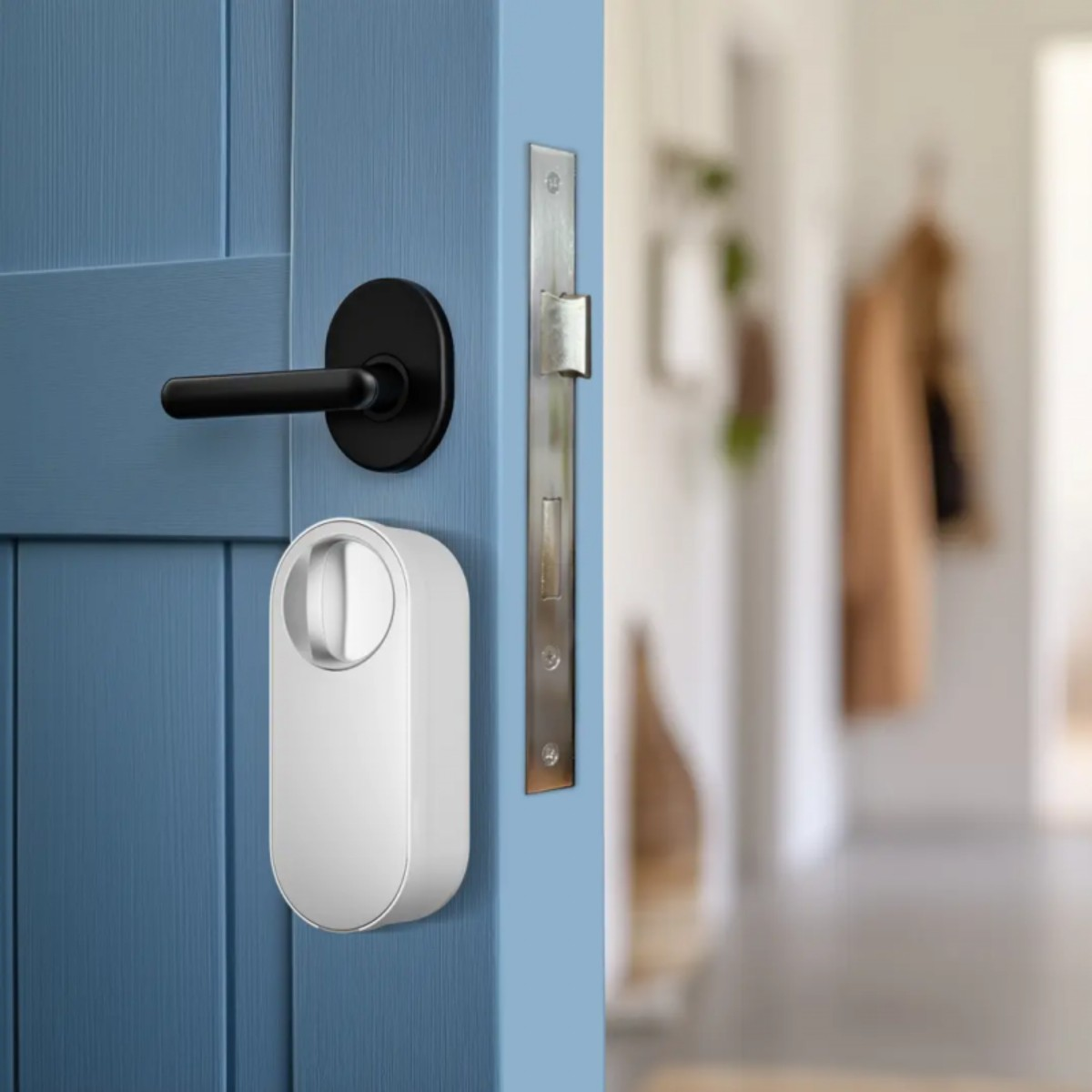 Eine blaue Tür mit schwarzem Griff und einem weißen Smart Lock. Dahinter ist ein Flur mit hängenden Mänteln leicht unscharf.