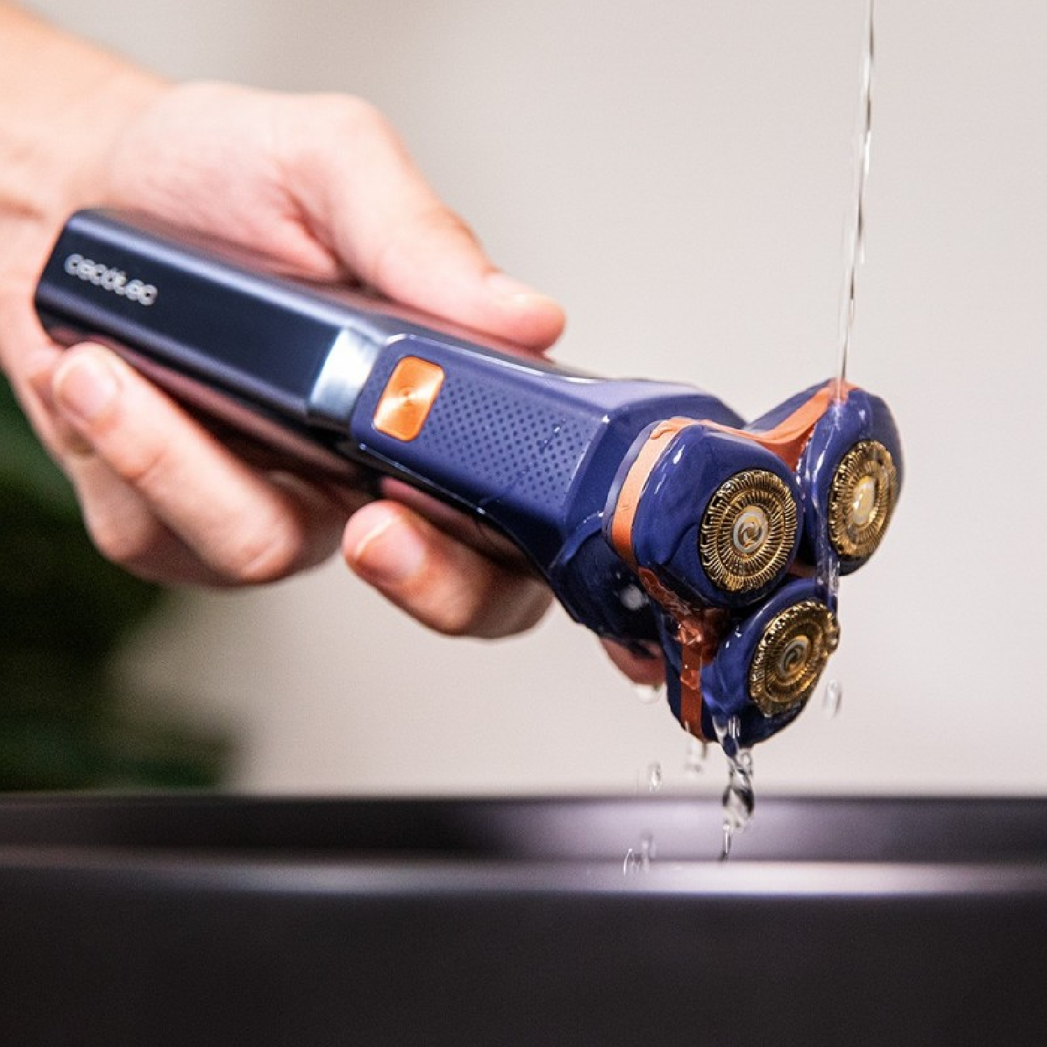 Un rasoio elettrico blu e arancione viene tenuto, l'acqua scorre sulle testine. Lo sfondo è sfocato, che mostra un bacino scuro.