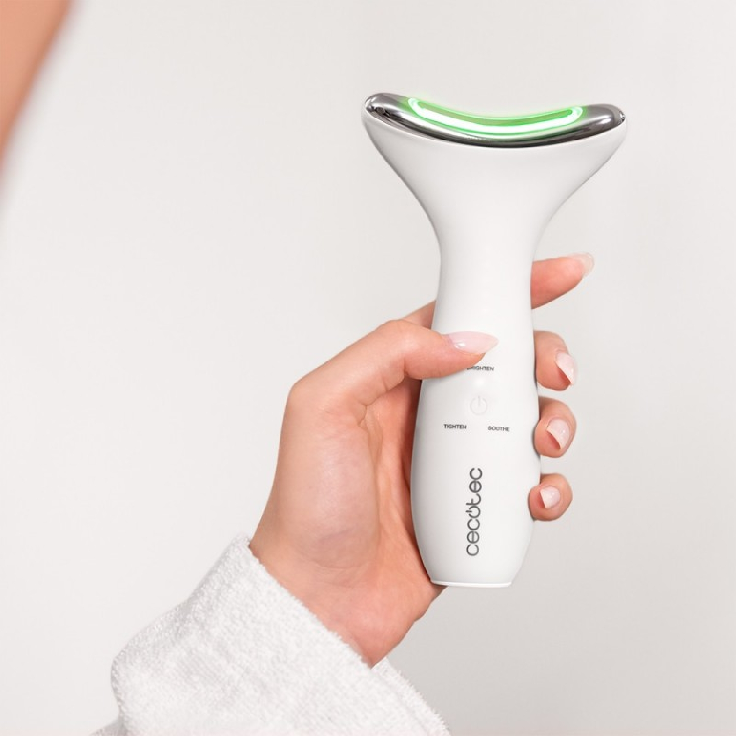Una mano sostiene un dispositivo de belleza blanco con una parte superior plateada. El dispositivo tiene luz verde y la marca 'cecotec' es visible.