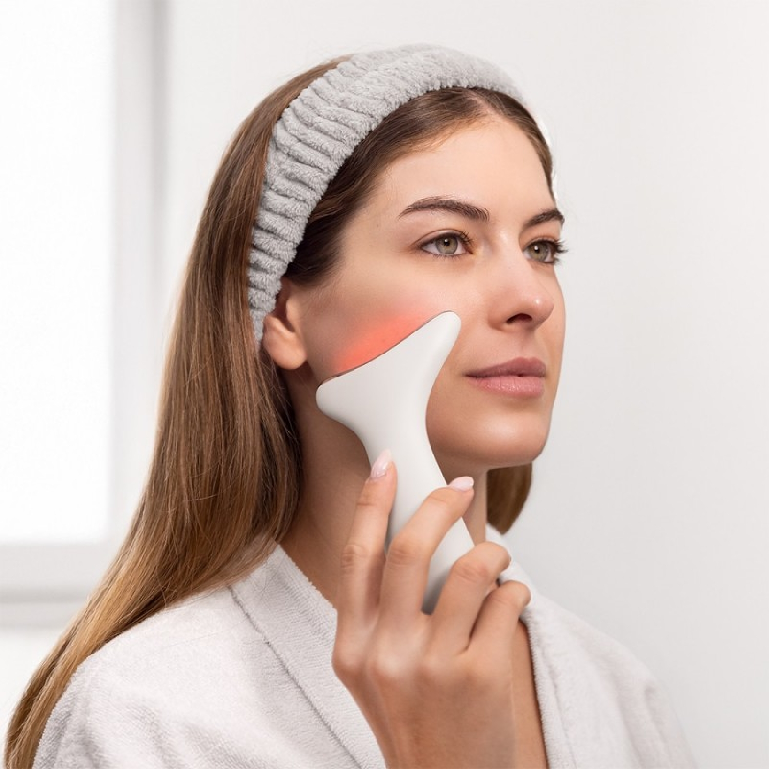 Mujer sosteniendo un dispositivo blanco para el cuidado de la piel en su rostro. Lleva una diadema gris y una bata blanca. Luz roja visible.