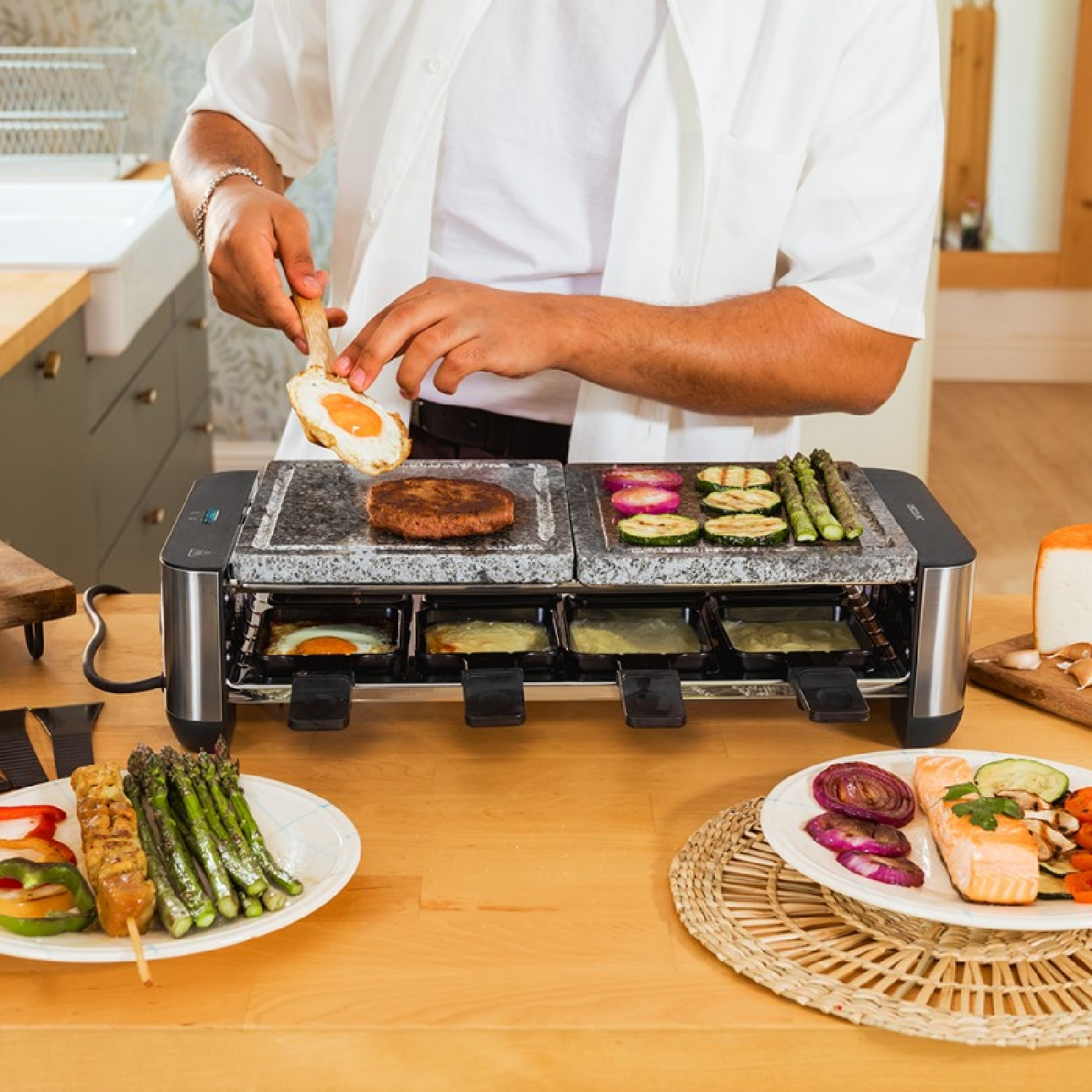 Osoba gotuje jedzenie na grillu do raclette. Są warzywa, mięso i jajka. Otoczenie to kuchnia z drewnianymi powierzchniami.