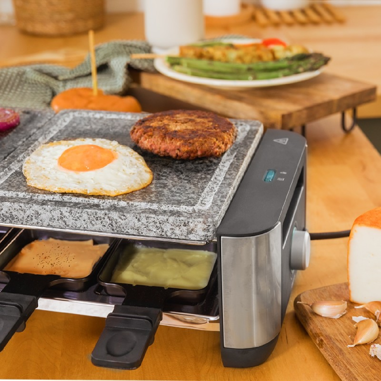 Grill do raclette z jajkiem, burgerem i serem. Szparagi i pomidor na talerzu. Czosnek i ser na drewnianej desce.