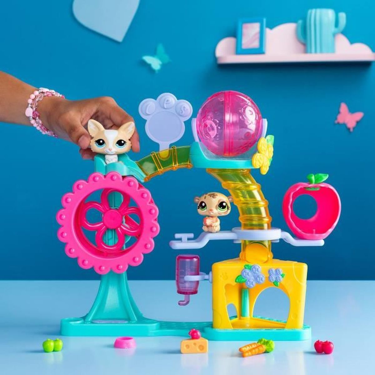 Un juego de juguetes para mascotas colorido con una rueda rosa, una bola de hámster y pequeñas figuras de animales. Una mano con pulsera en la parte superior.