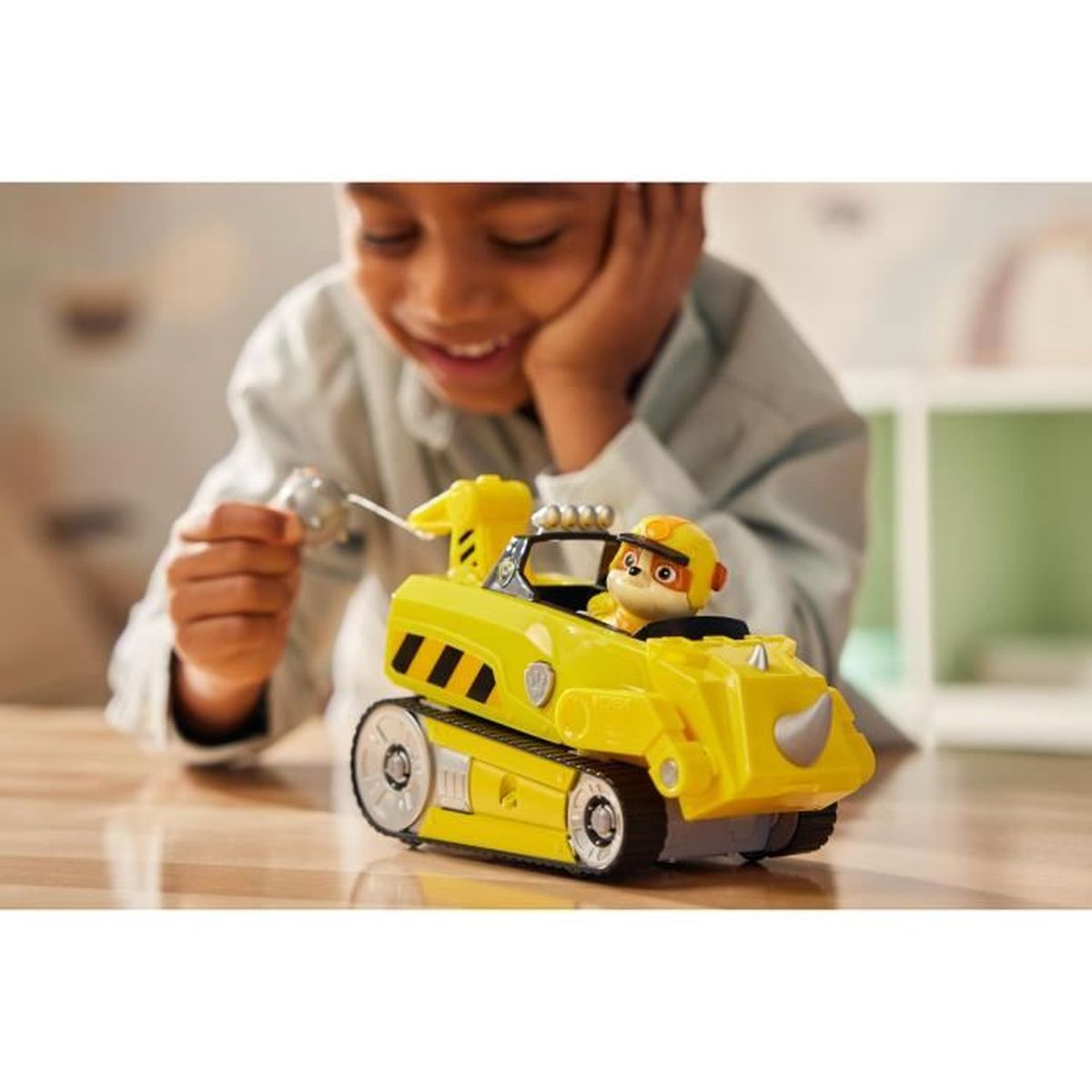 Ein Junge lächelt, während er ein Spielzeug hält. Ein gelbes Spielzeugauto mit einer Hundefigur steht auf einem Holztisch.