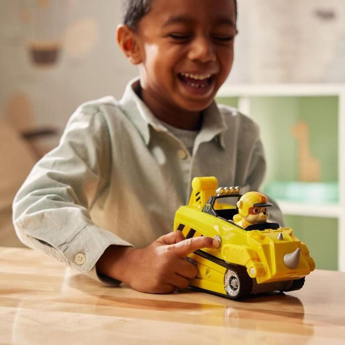 Ein lächelnder Junge zeigt auf ein gelbes Spielzeugauto mit einer Figur darin. Das Spielzeug steht auf einem Holztisch.