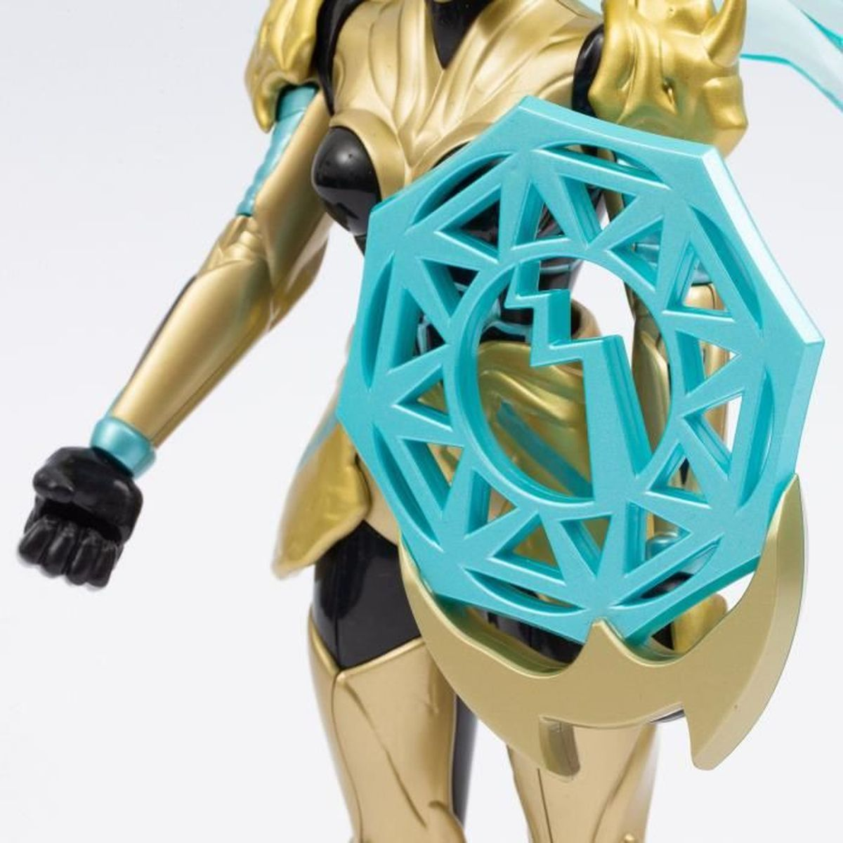 Nahaufnahme einer Figur mit goldener Rüstung, die einen türkisfarbenen Schild hält. Schwarze Handschuhe. Der Schild hat ein geometrisches Design.