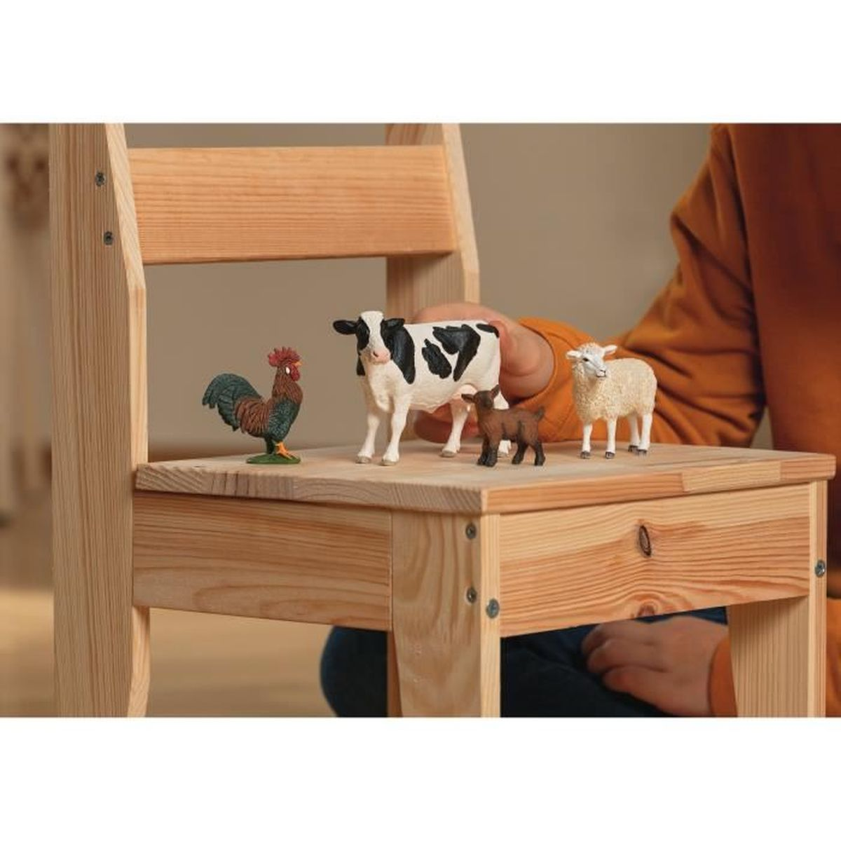 La main d'un enfant place des jouets d'animaux de ferme sur une chaise en bois. Un coq, une vache, une chèvre et un mouton sont visibles.