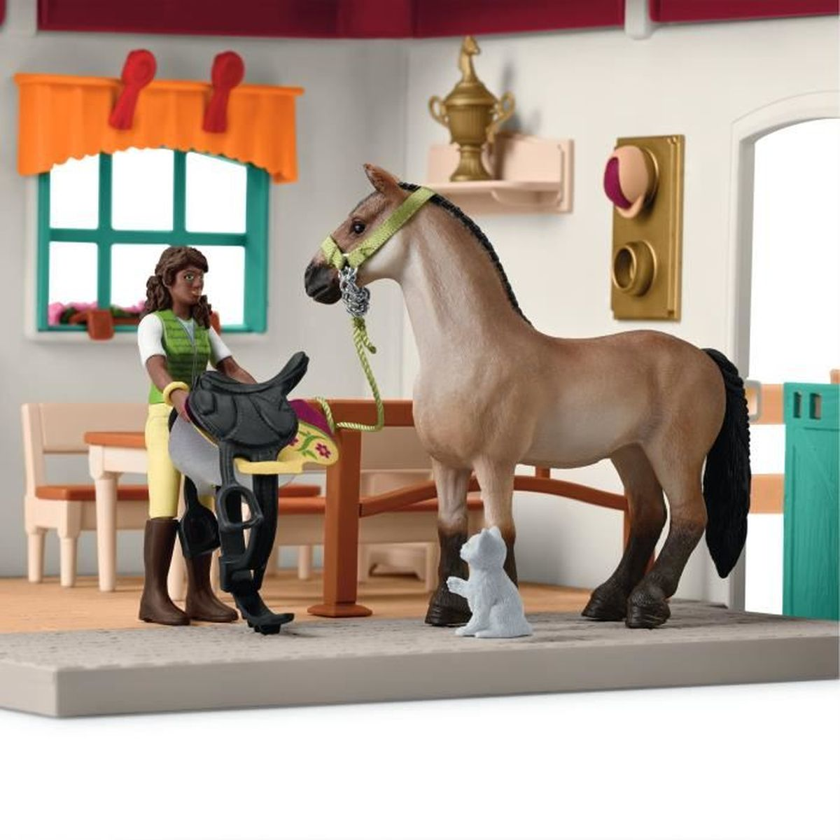 Un cavallo e un cavaliere giocattolo con una sella in un ambiente da stalla. Un gatto è di fronte al cavallo. La scena include una finestra e un trofeo.