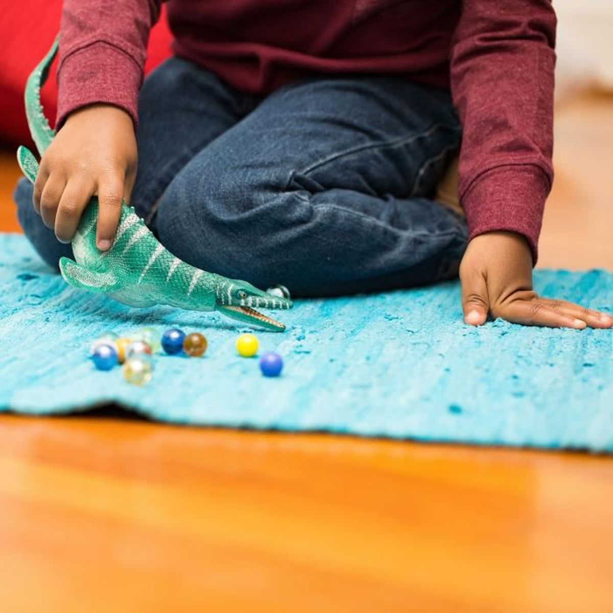 Dziecko w czerwonej koszuli i dżinsach bawi się zabawką dinozaura na niebieskim dywanie. Rozrzucone są kulki w różnych kolorach.