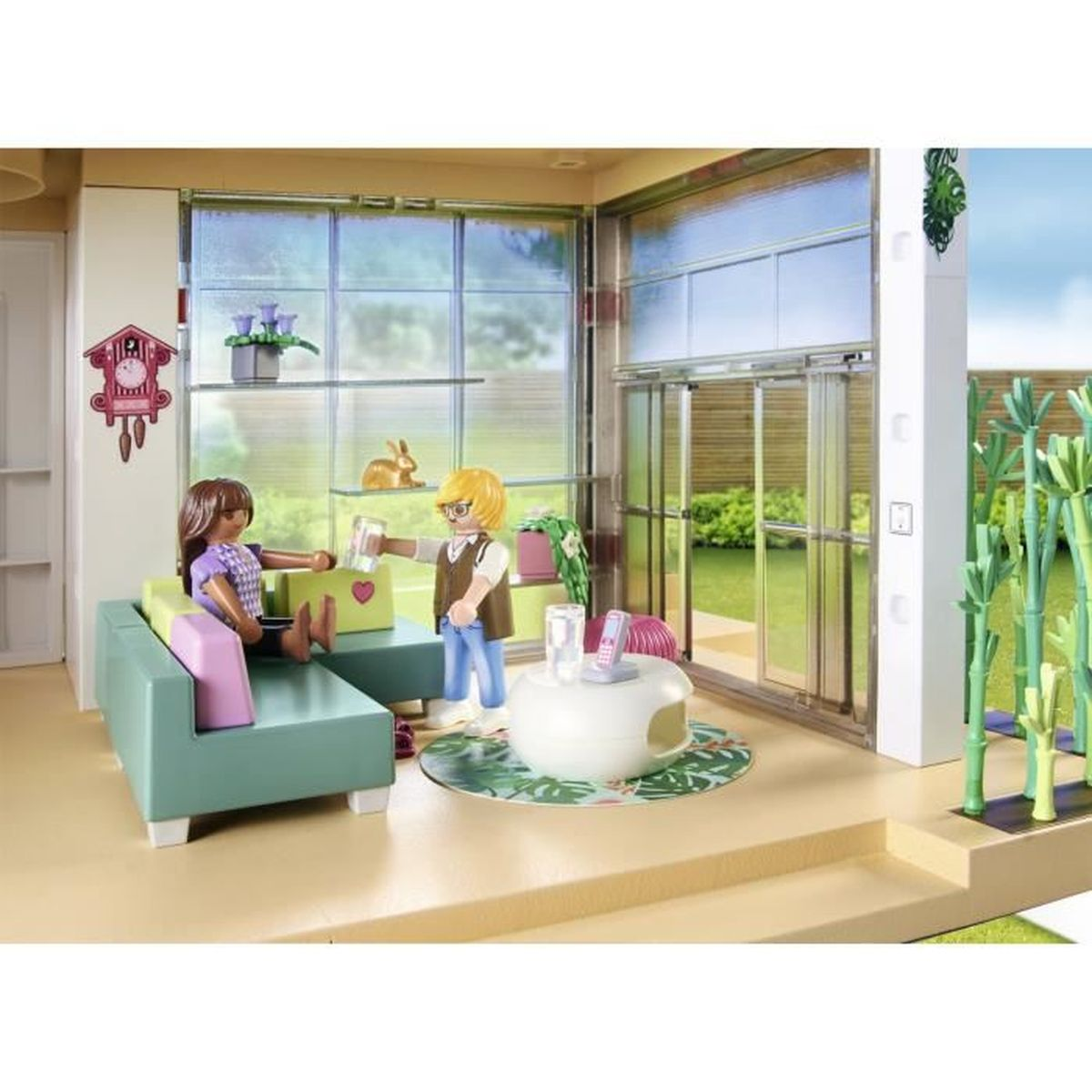 Zwei Figuren in einem Spielzeughaus, eine bietet ein Getränk an. Sofa, Tisch und Dekoration sind sichtbar. Durch das Glas ist ein Garten zu sehen.