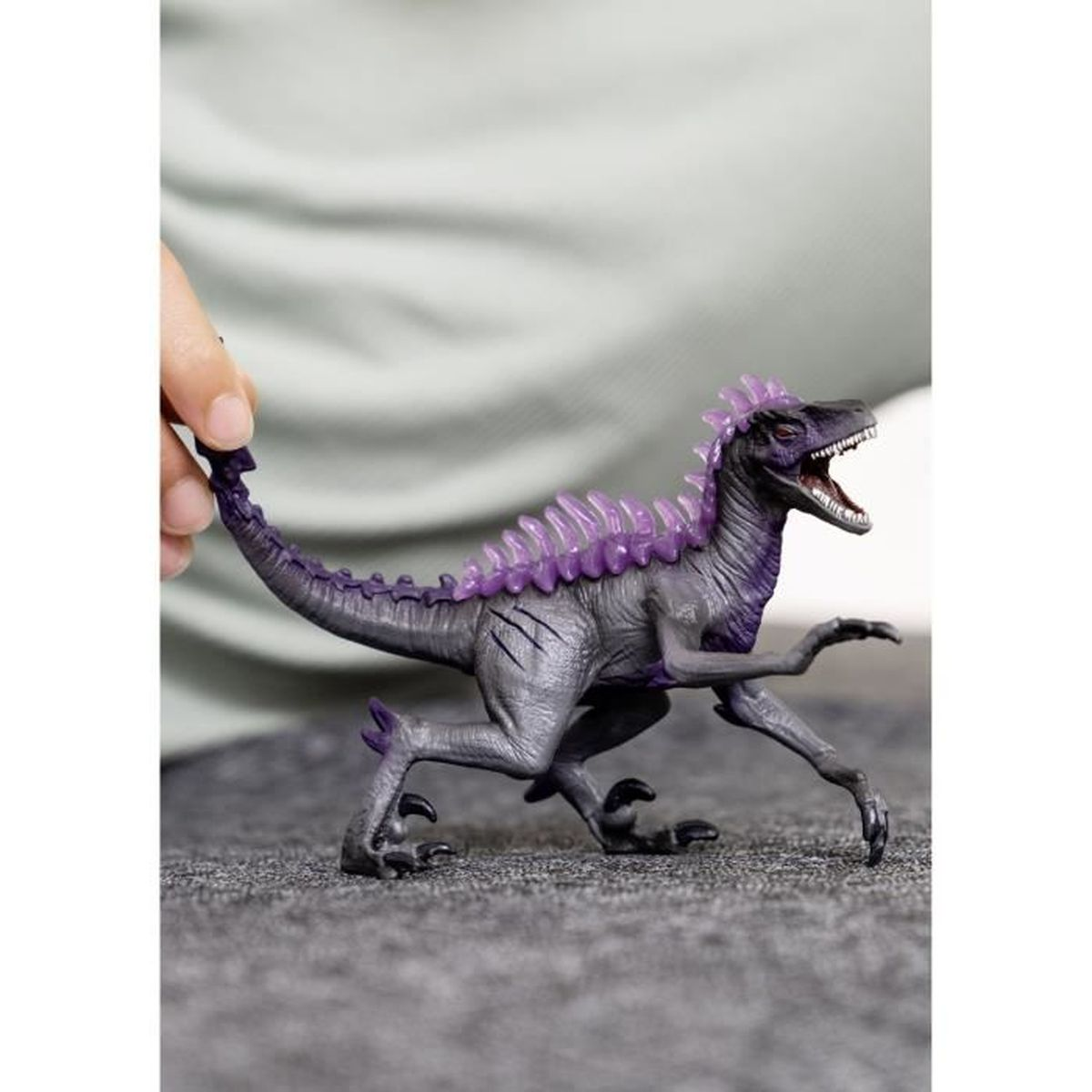 Ein graues Dinosaurierspielzeug mit lila Stacheln und Kamm wird von einer Hand gehalten. Das Spielzeug befindet sich auf einer grauen Oberfläche mit hellem Hintergrund.
