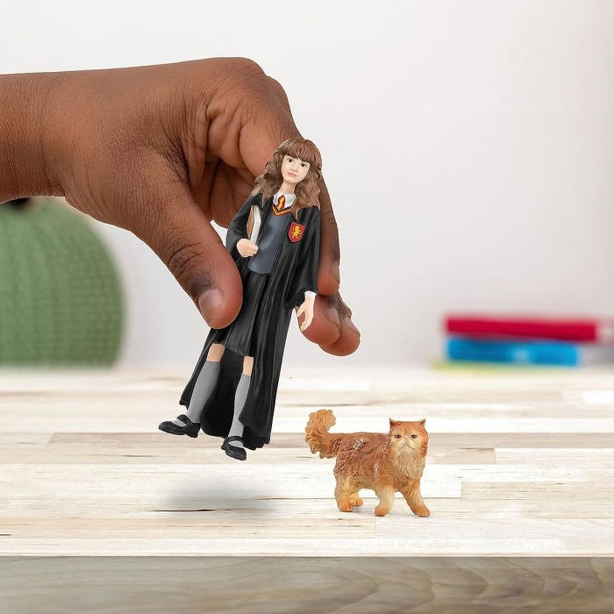 Una mano tiene una statuetta di Hermione Granger, accanto a una statuetta di gatto. Entrambe le statuette sono su una superficie di legno. Alcune matite sullo sfondo.