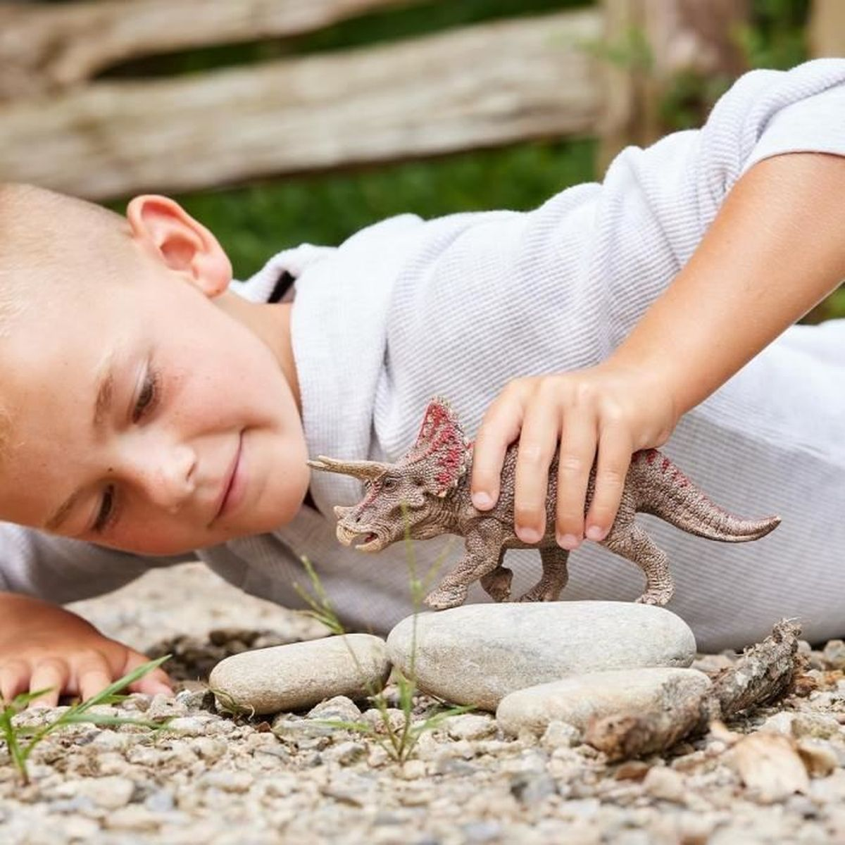 Chłopiec ogląda zabawkowego dinozaura. Dinozaur stoi na kamieniu wśród żwiru. W tle drewniany płot i zielona trawa.