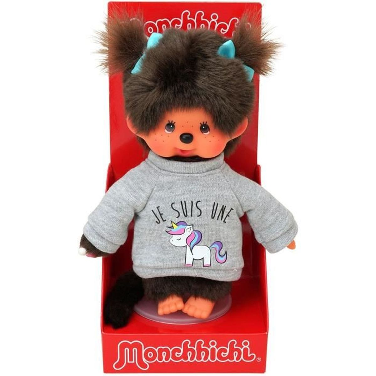 Lalka Monchhichi w szarym swetrze z jednorożcem. Ma brązowe futro, niebieskie kokardy i jest w czerwonym pudełku.